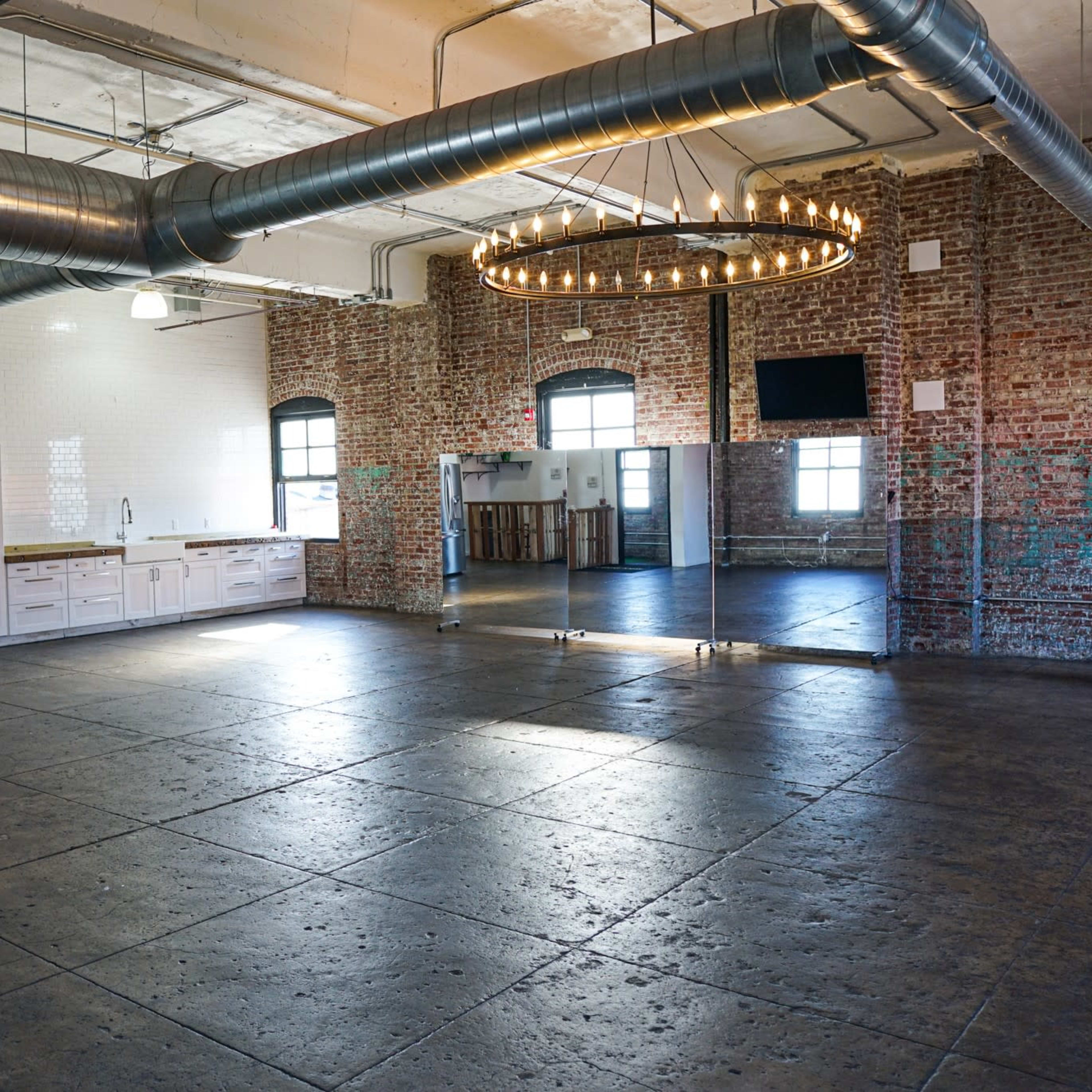 An open, industrial-style space features exposed brick walls, a kitchen area, and large mirrors along one side.
