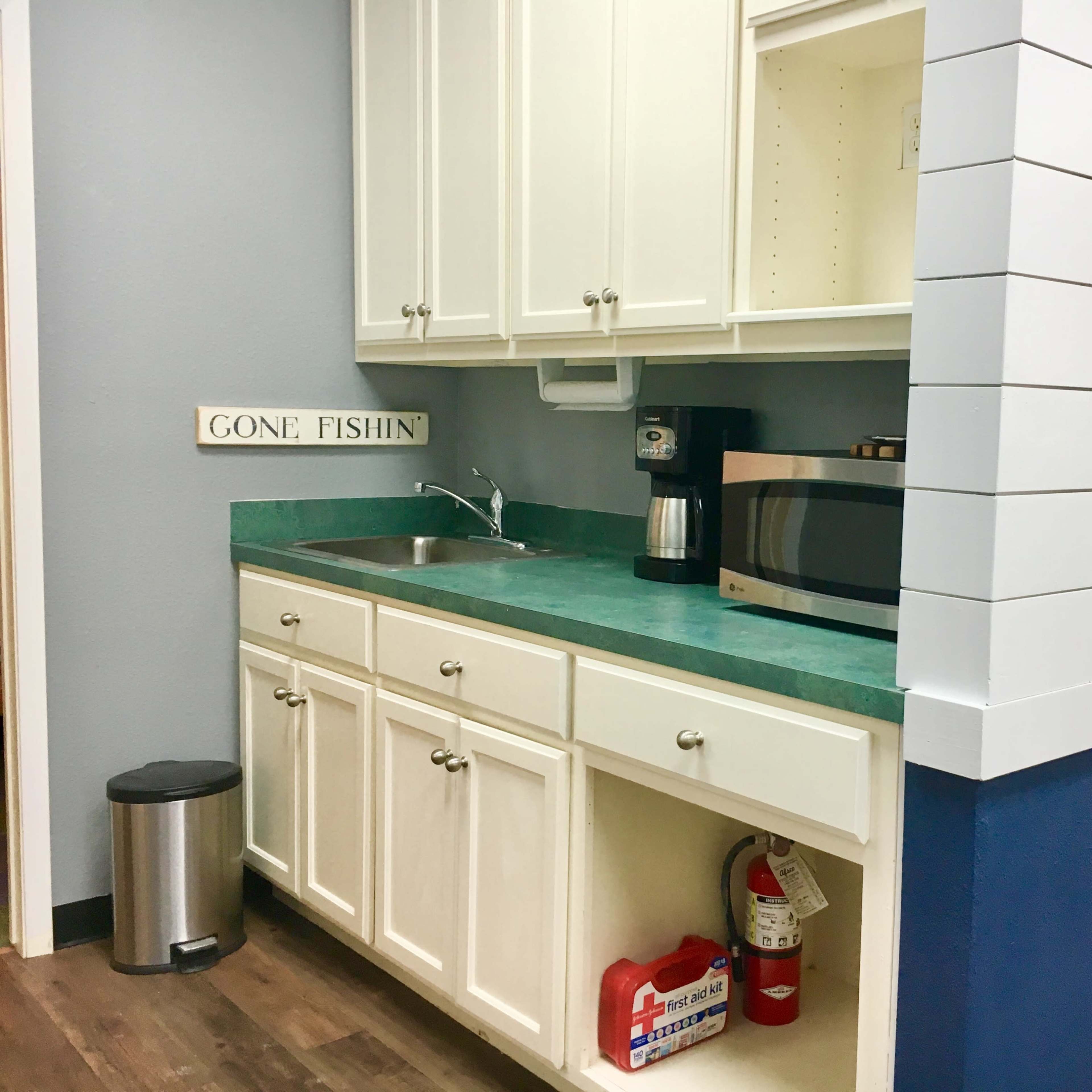 A kitchen area with white cabinets, a green countertop, a sink, a coffee maker, a microwave, and a wall painted in blue, alongside a sign that reads "GONE FISHIN'."