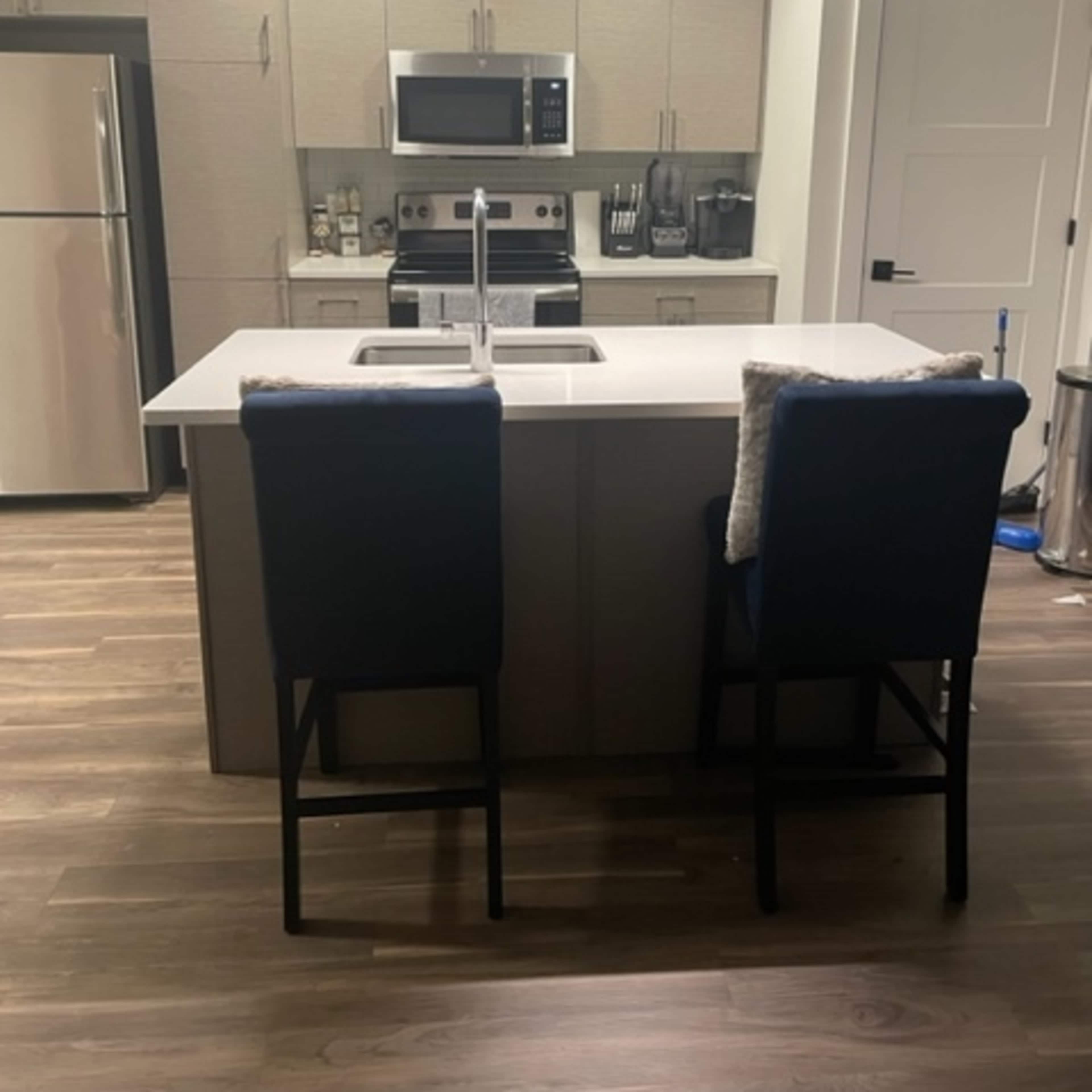 A modern kitchen with a central island featuring two bar stools, stainless steel appliances, and light wood flooring.
