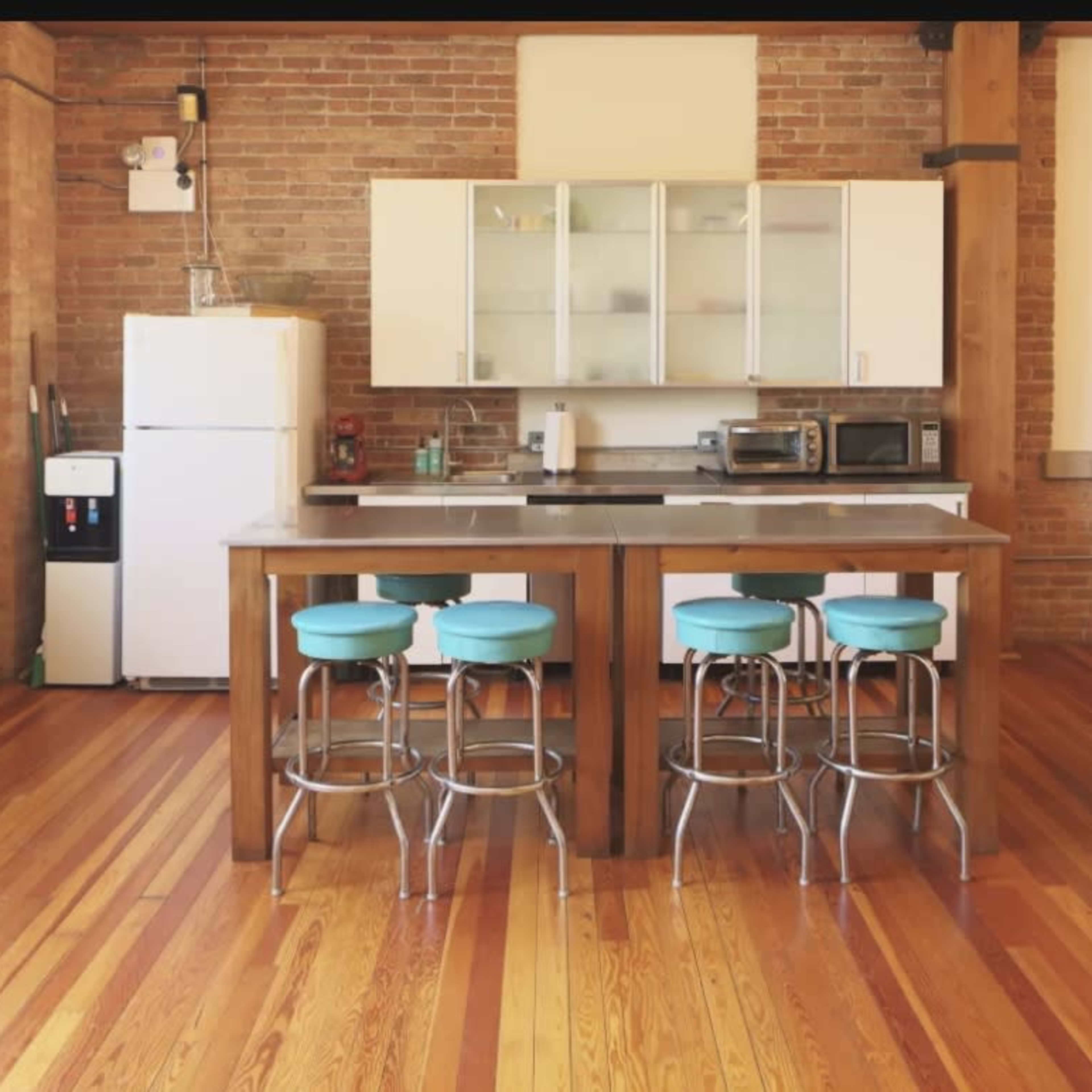 A kitchen area with a wooden island and three teal bar stools, complemented by a refrigerator, microwave, and an open living space with brick walls and hardwood floors.