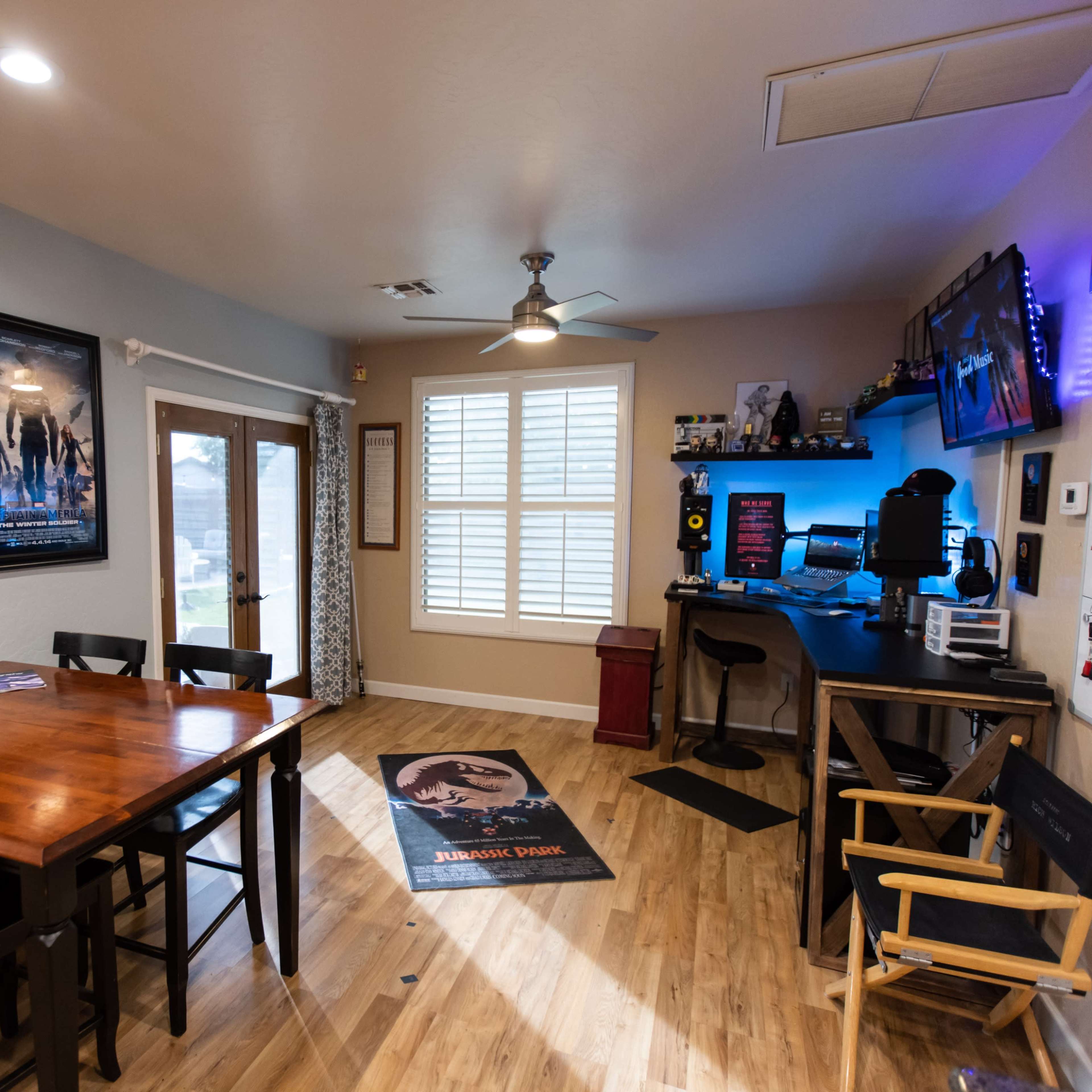 A modern room with a wooden dining table, film posters on the walls, a desk setup with a computer, and a door leading outside.