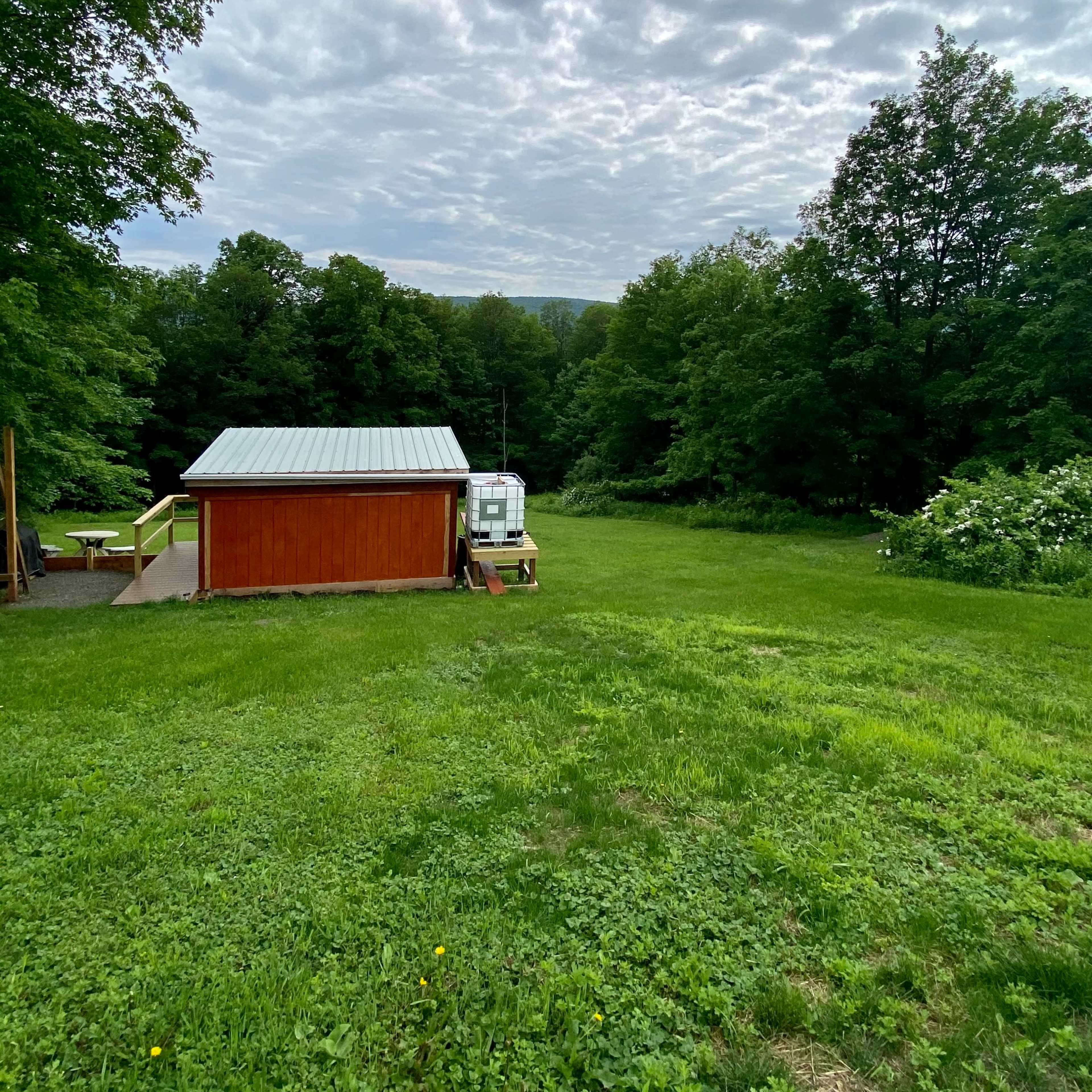 A small wooden shed with a metal roof is situated in a green grassy area surrounded by trees under a cloudy sky.
