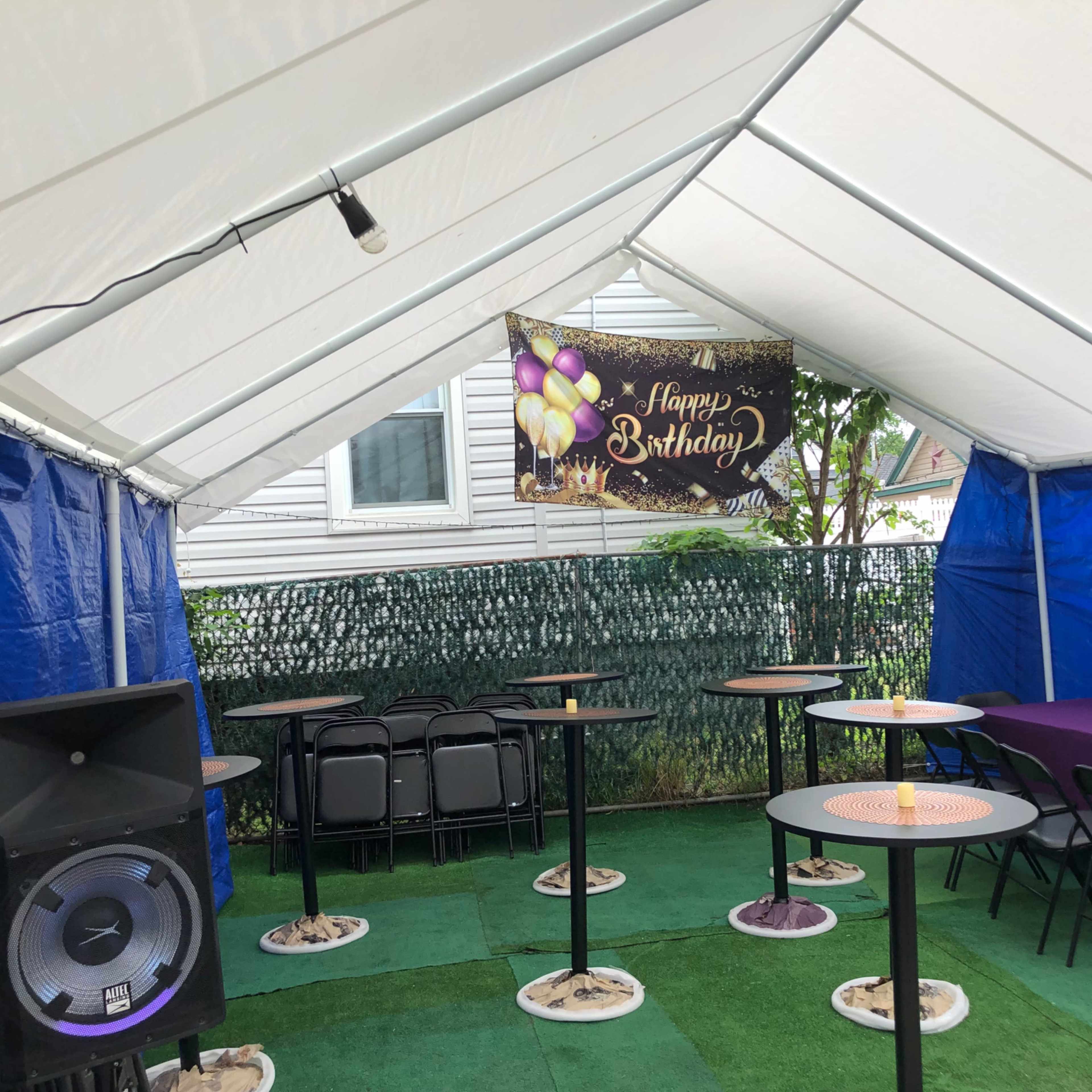 A party tent is set up with tables and chairs, featuring a "Happy Birthday" banner and a speaker in one corner.