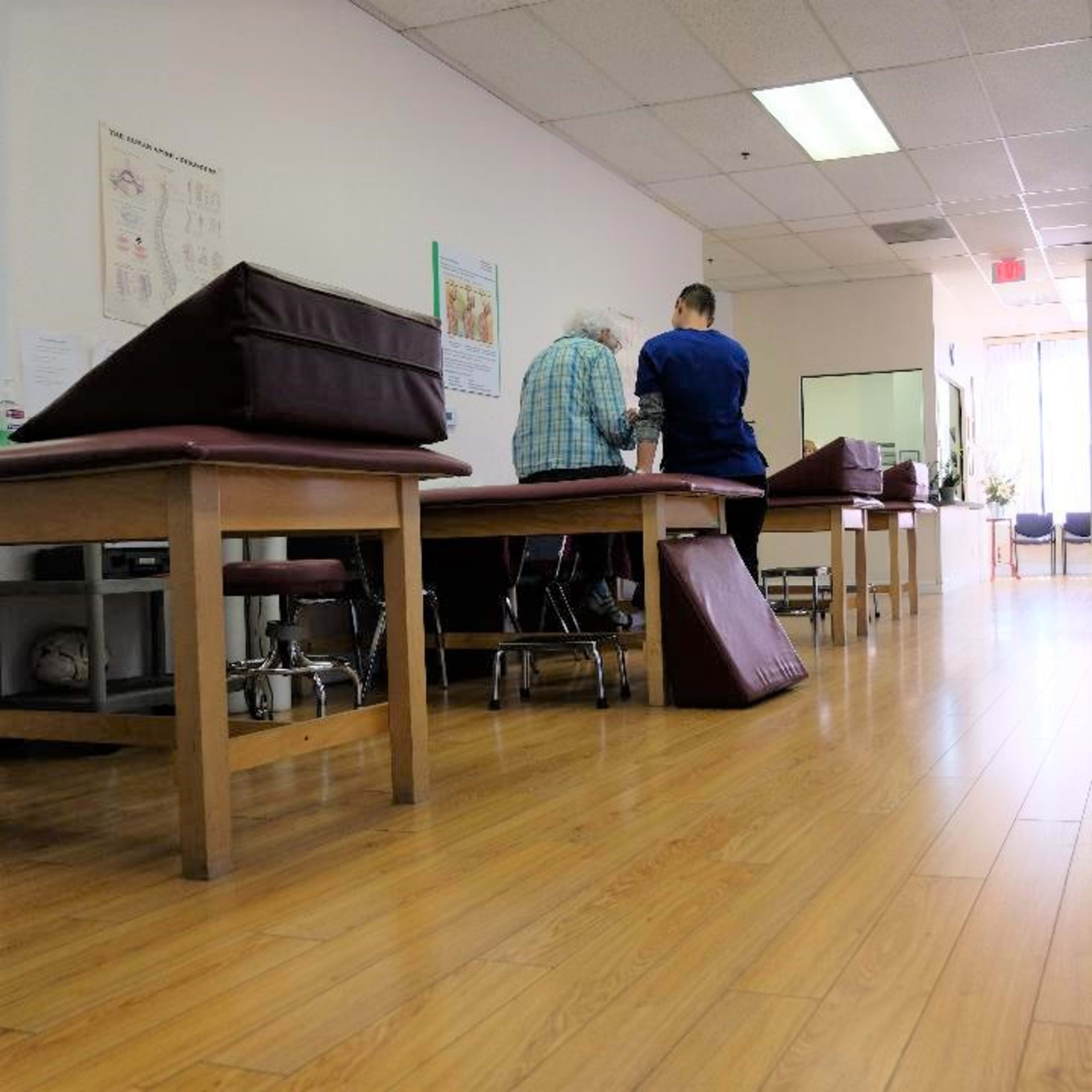 A physical therapy room features several treatment tables, a patient and therapist engaged in an activity, and equipment along the side.