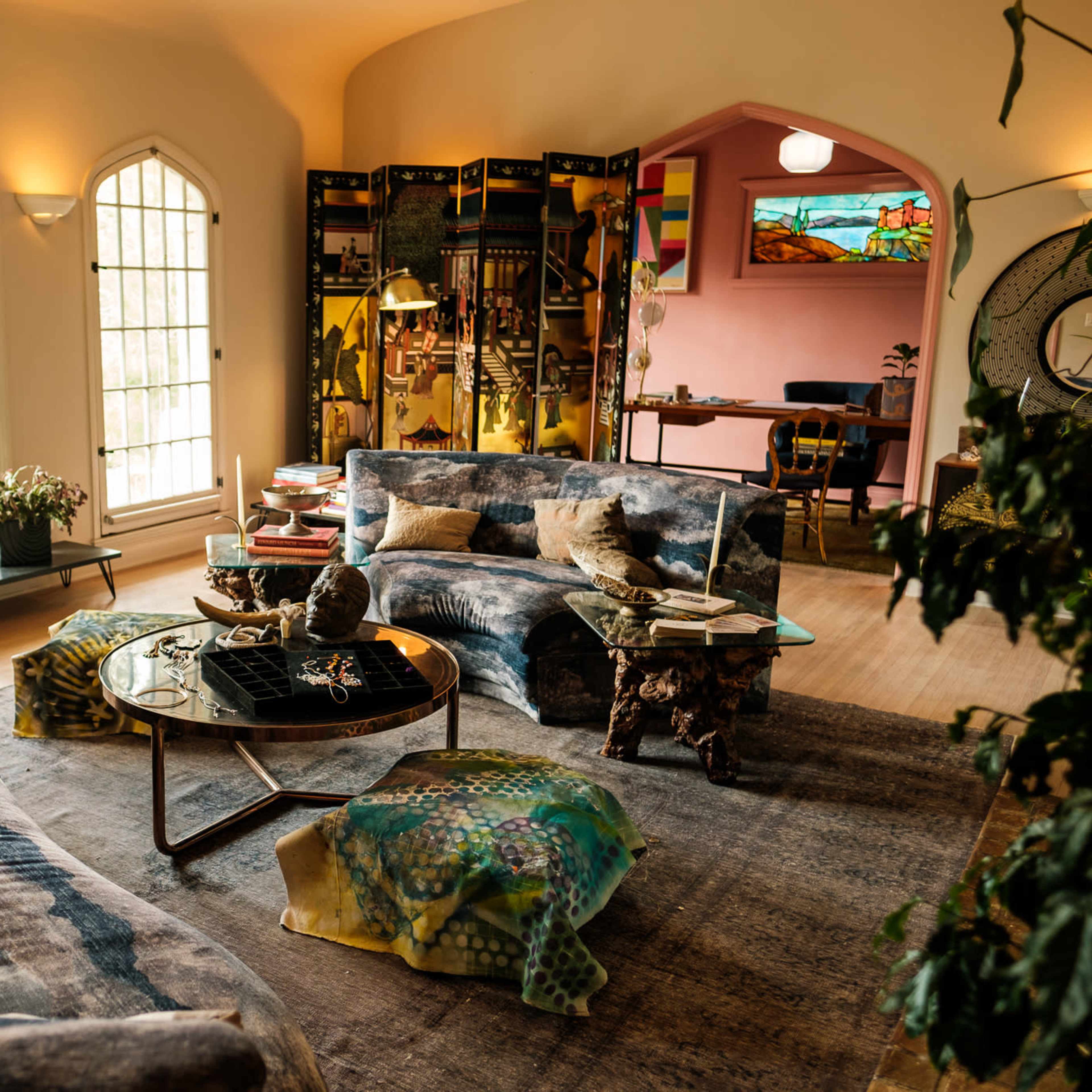 A well-lit living room with curved couches, a coffee table, and various plants, framed by large windows and colorful decor.