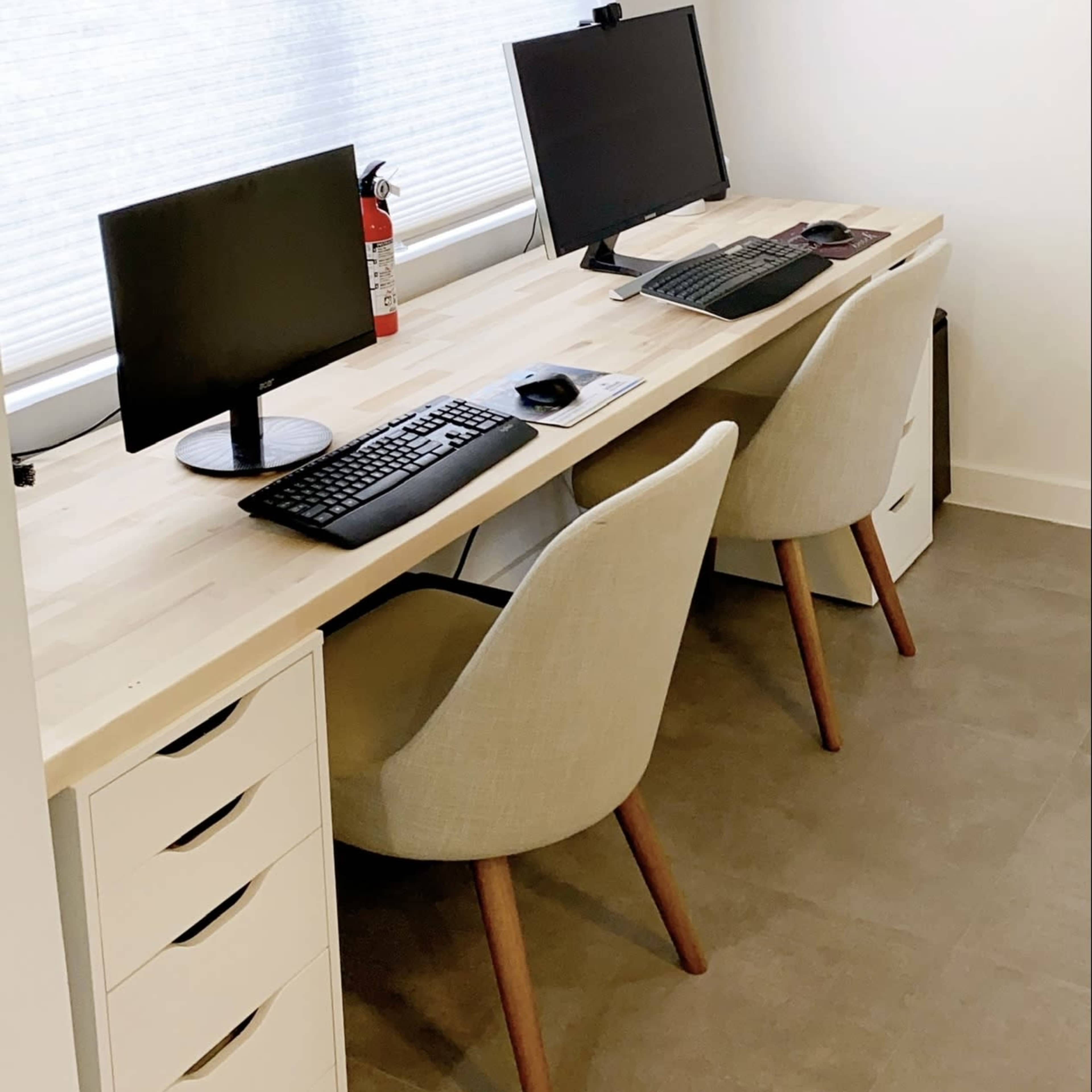 A minimalist workspace features two computers on a shared desk, paired with light-colored chairs and a window with sheer blinds.
