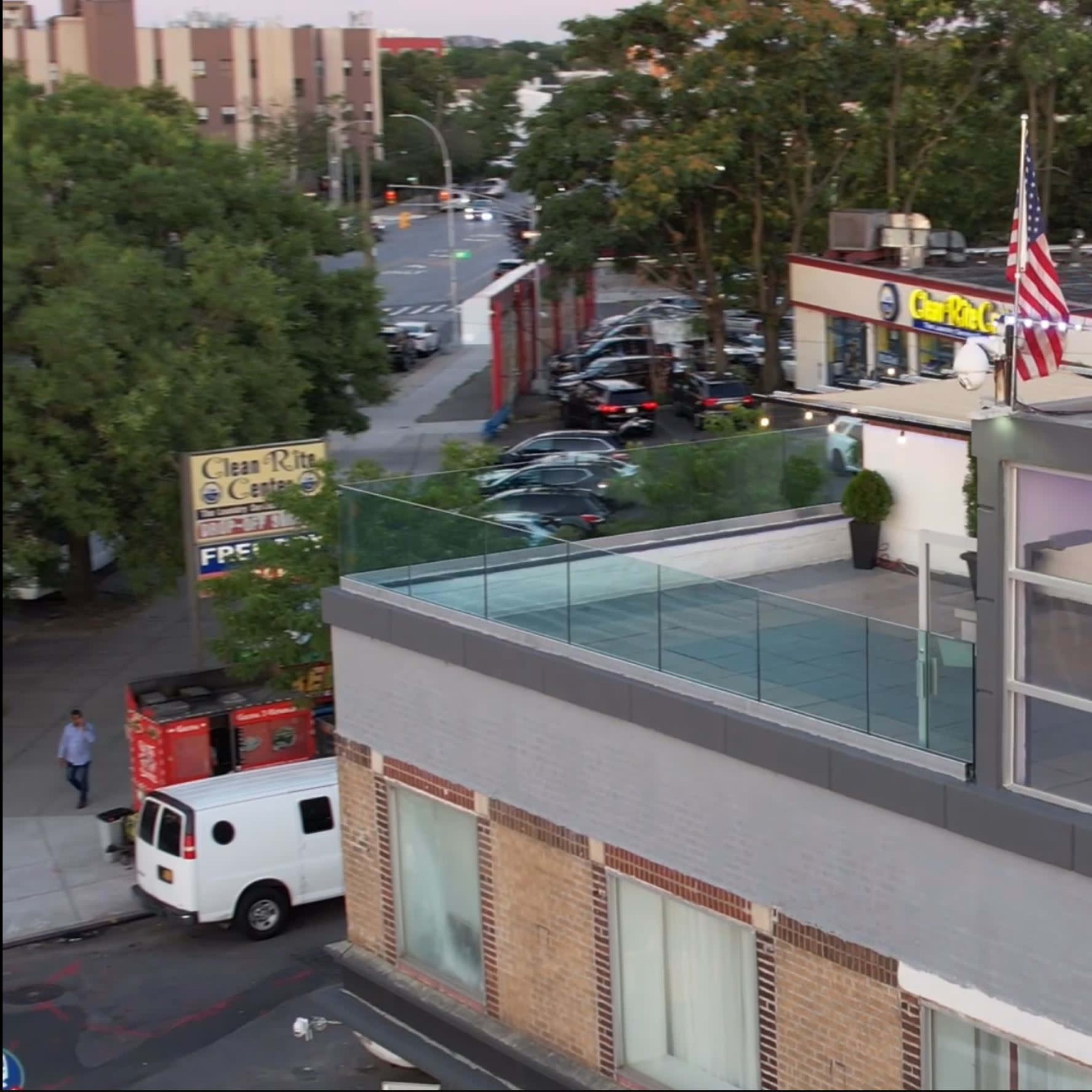 A rooftop terrace with glass railing overlooks a street lined with parked cars and trees, featuring an American flag on a pole.