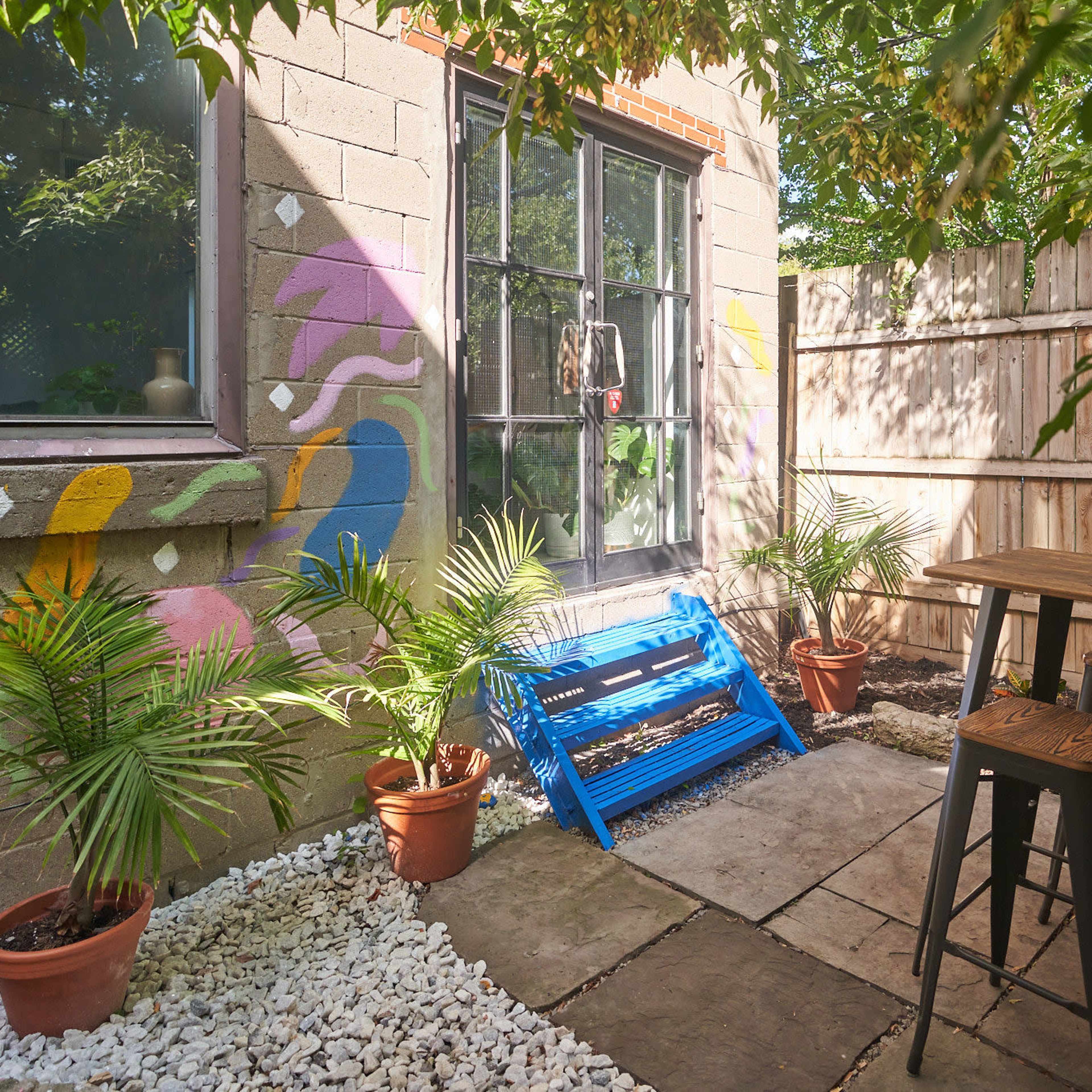 A small outdoor patio features a blue lounge chair, a wooden table with stools, and potted plants surrounded by a decorative stone pathway and colorful wall art.