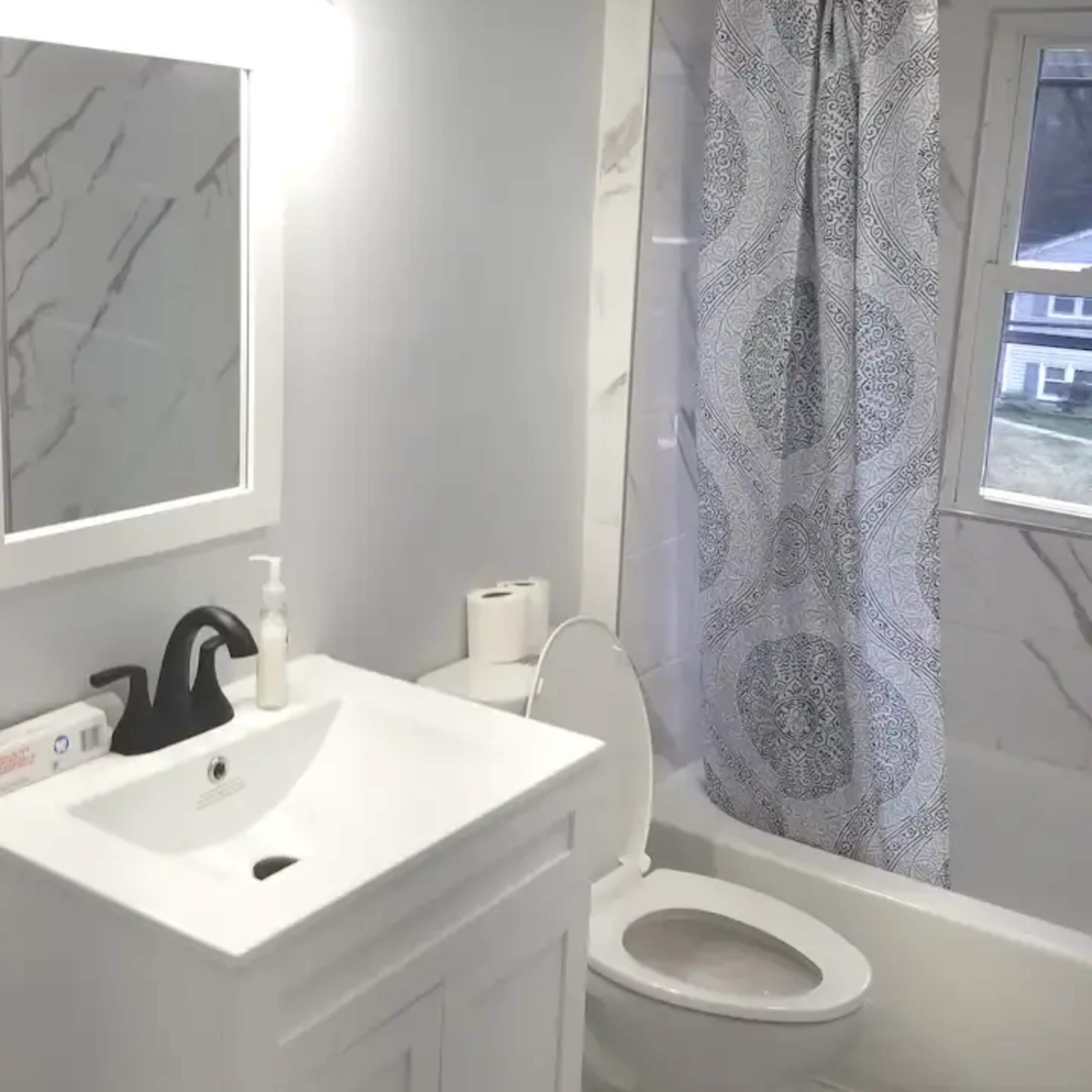 A small bathroom featuring a white vanity with a sink, a mirror, a black faucet, a toilet, and a bathtub with a patterned shower curtain.