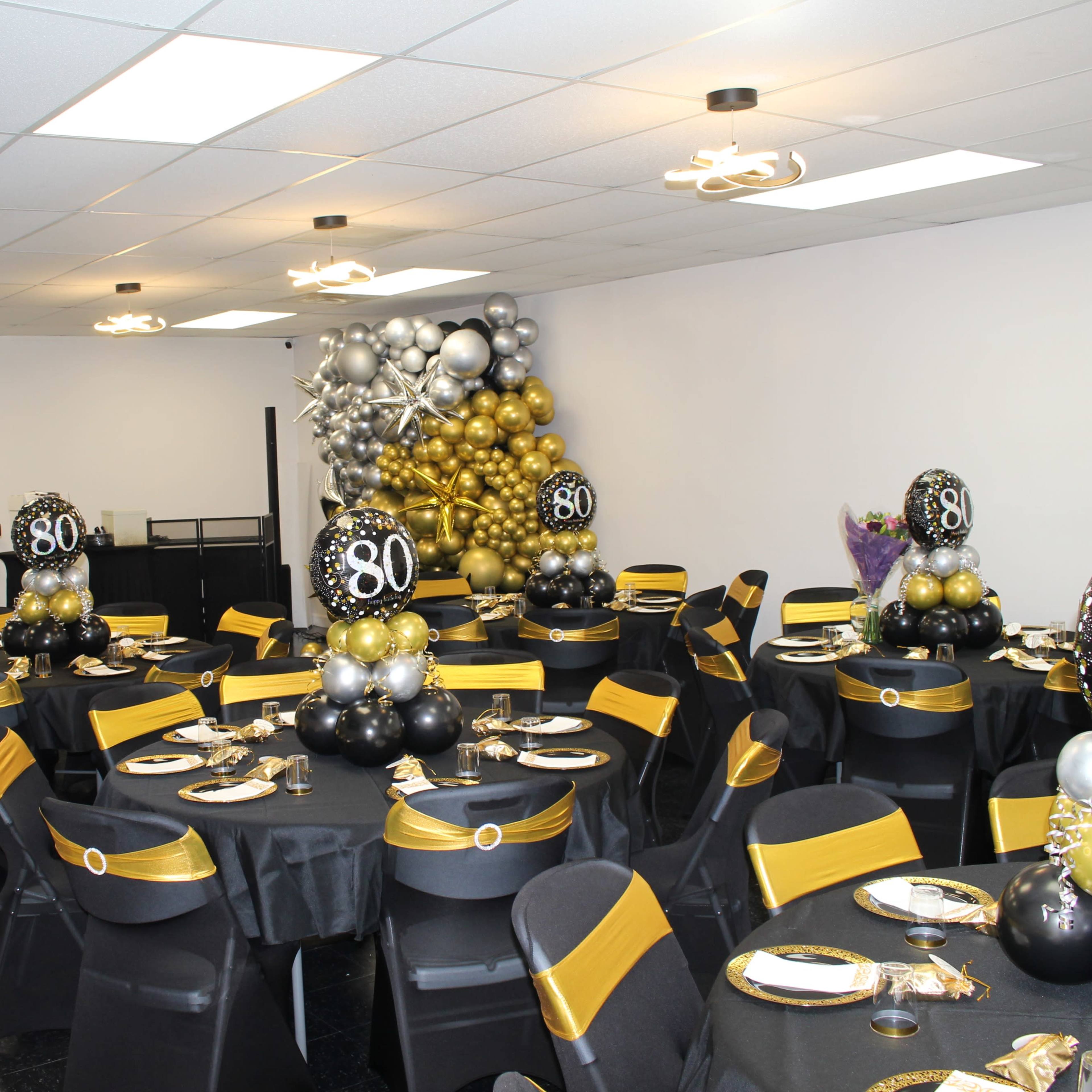 A decorated banquet room features black and gold table settings with balloon centerpieces celebrating an 80th birthday.