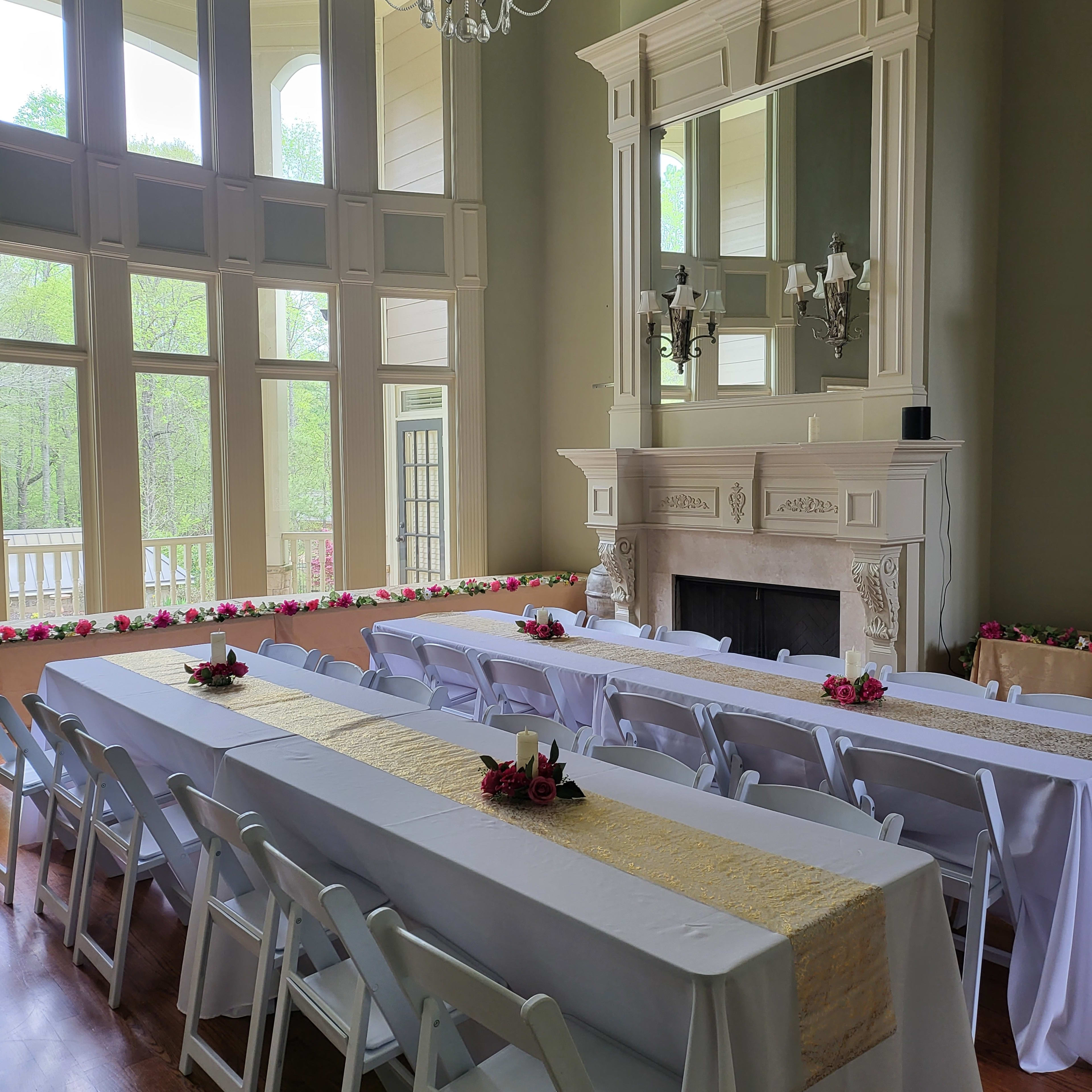 A spacious room with large windows, featuring multiple long tables set with white tablecloths and decorative centerpieces, alongside a fireplace and a chandelier above.