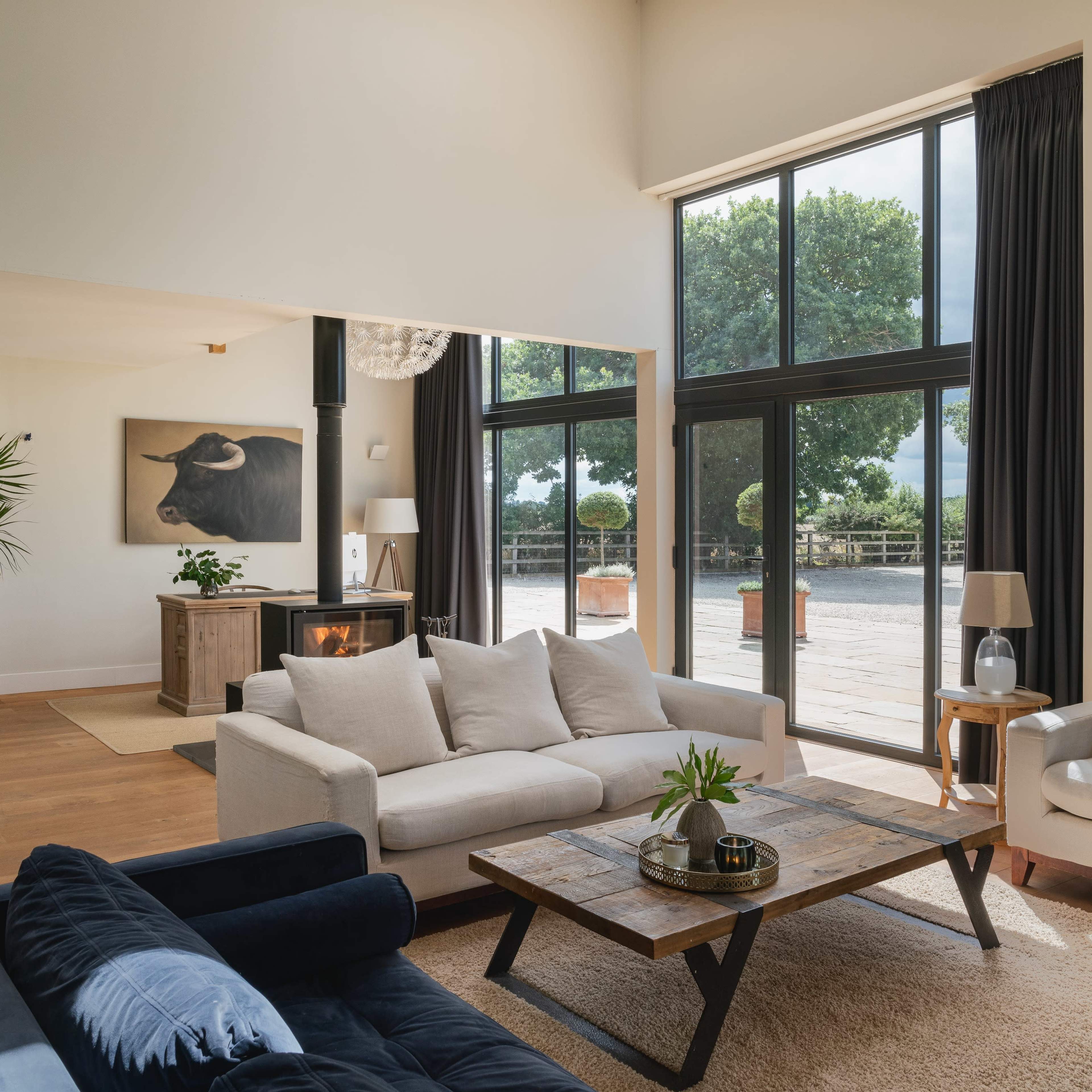 A spacious living room featuring a light-colored sofa, a dark blue couch, a wooden coffee table, and large windows with views of greenery outside.