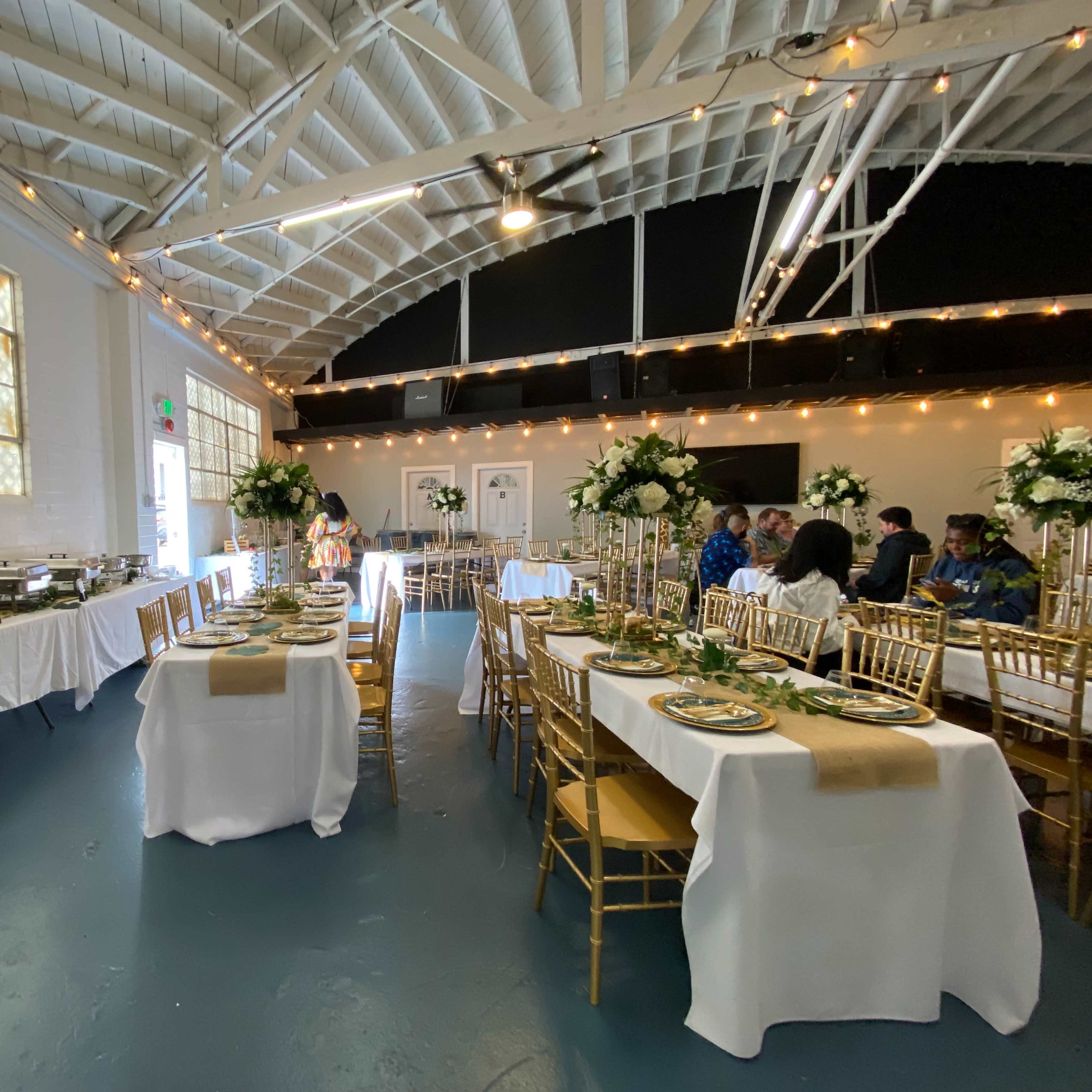 A large event space is arranged with gold chairs and tables set for a formal gathering, featuring buffet stations and floral decorations.