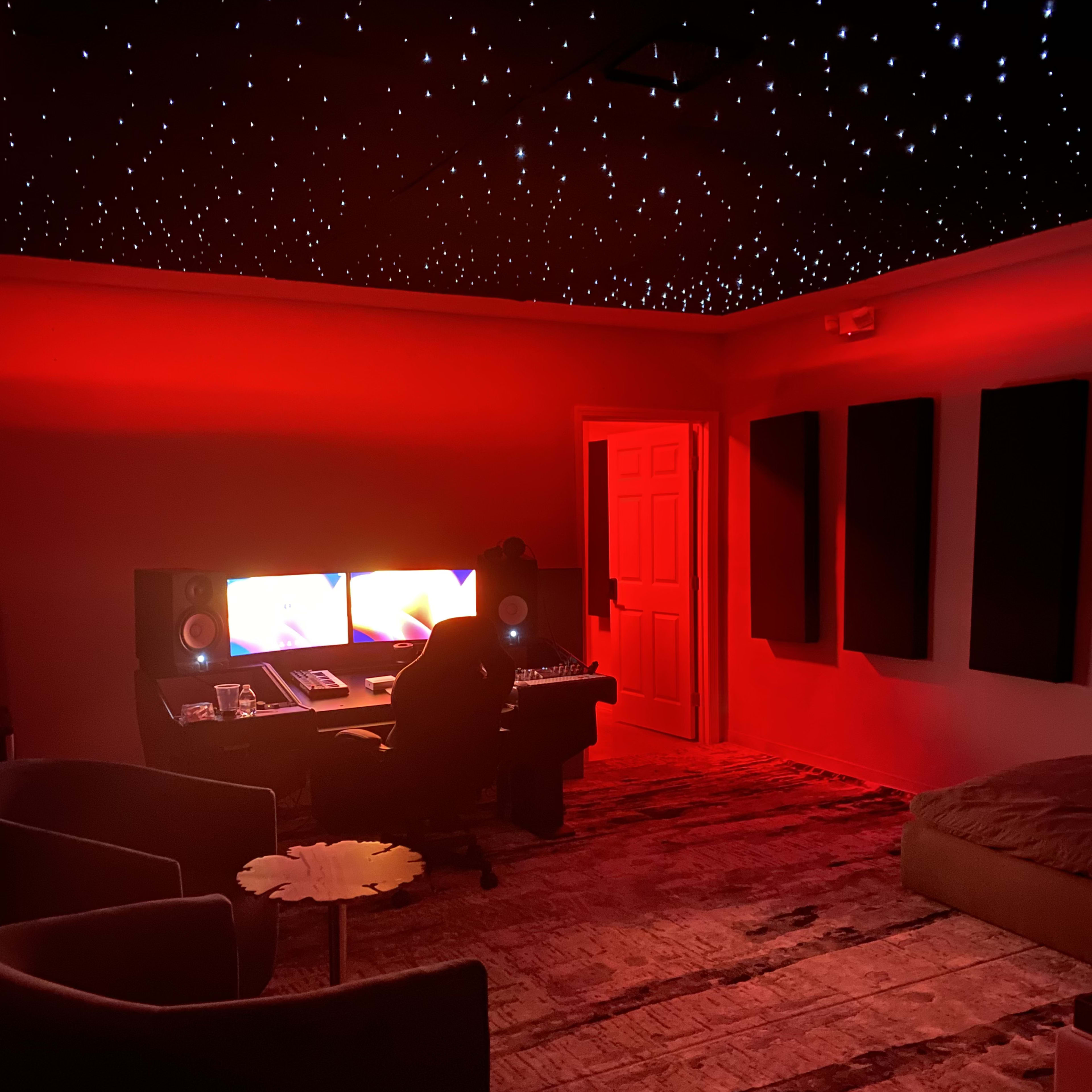 A dimly lit music studio features red lighting, a starry ceiling, and a central workstation with dual monitors.