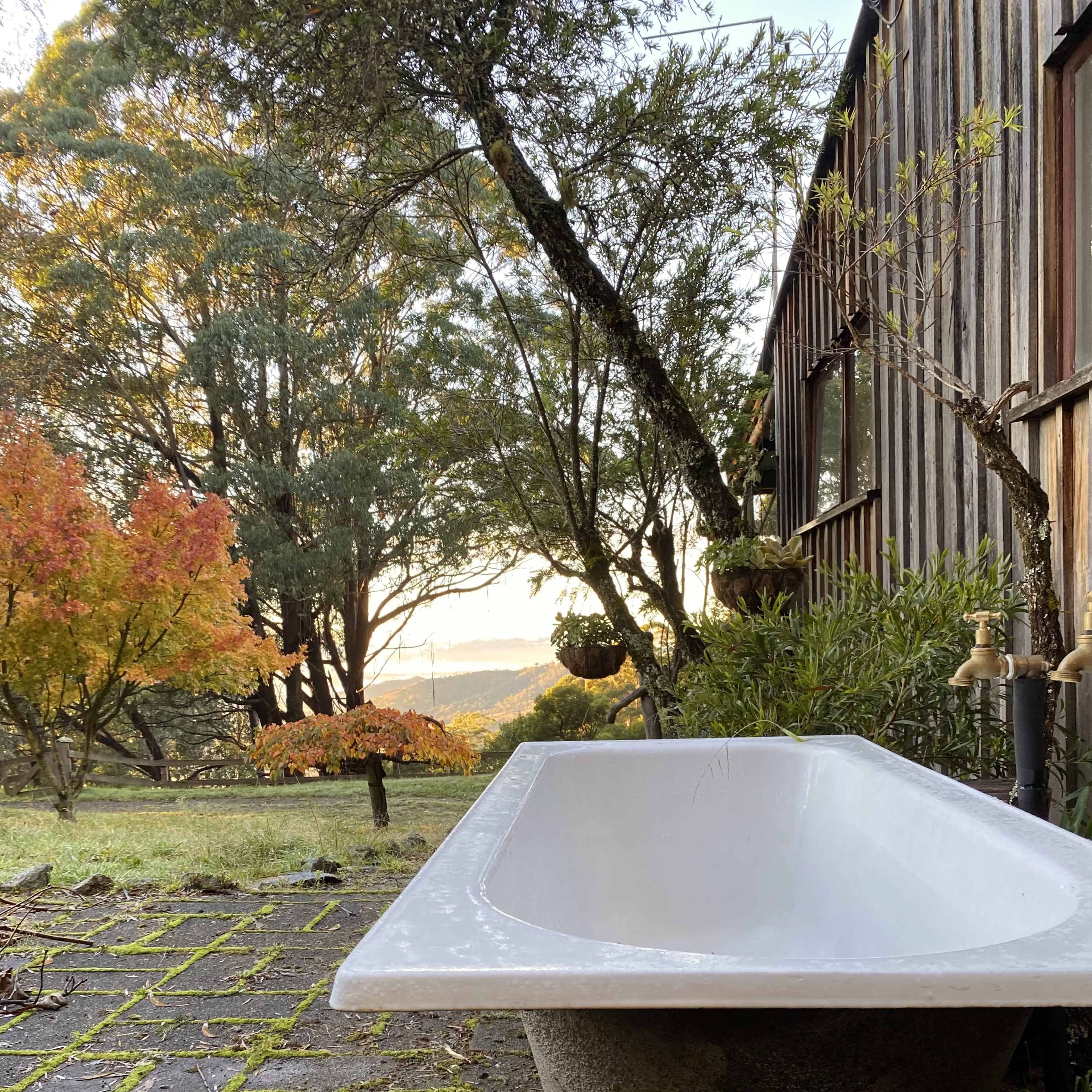 A freestanding bathtub is positioned on a stone patio, surrounded by trees and greenery, with colorful autumn foliage in the background.