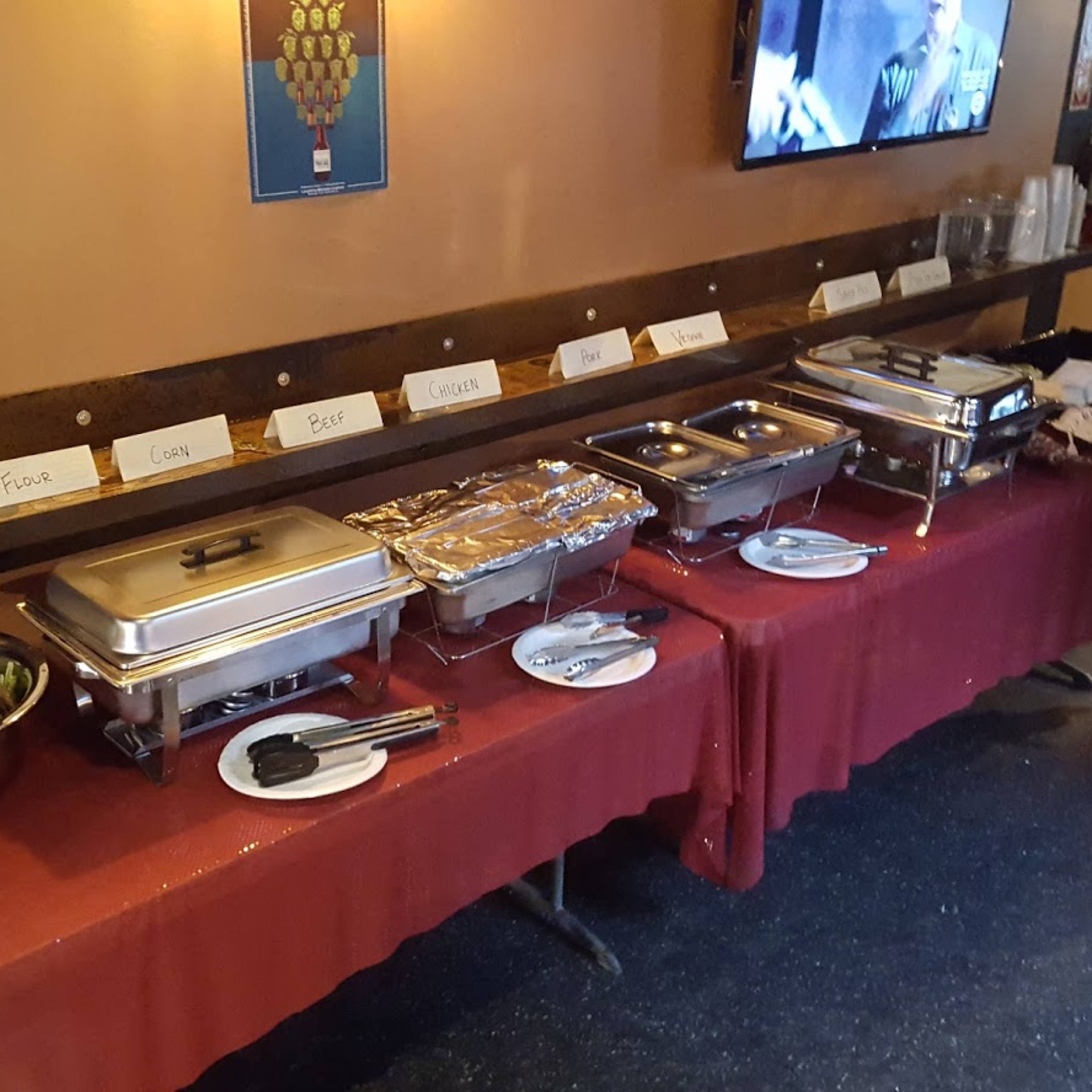 A buffet setup displays multiple metal chafing dishes and a large bowl of salad on a red tablecloth against a wall with a TV above.