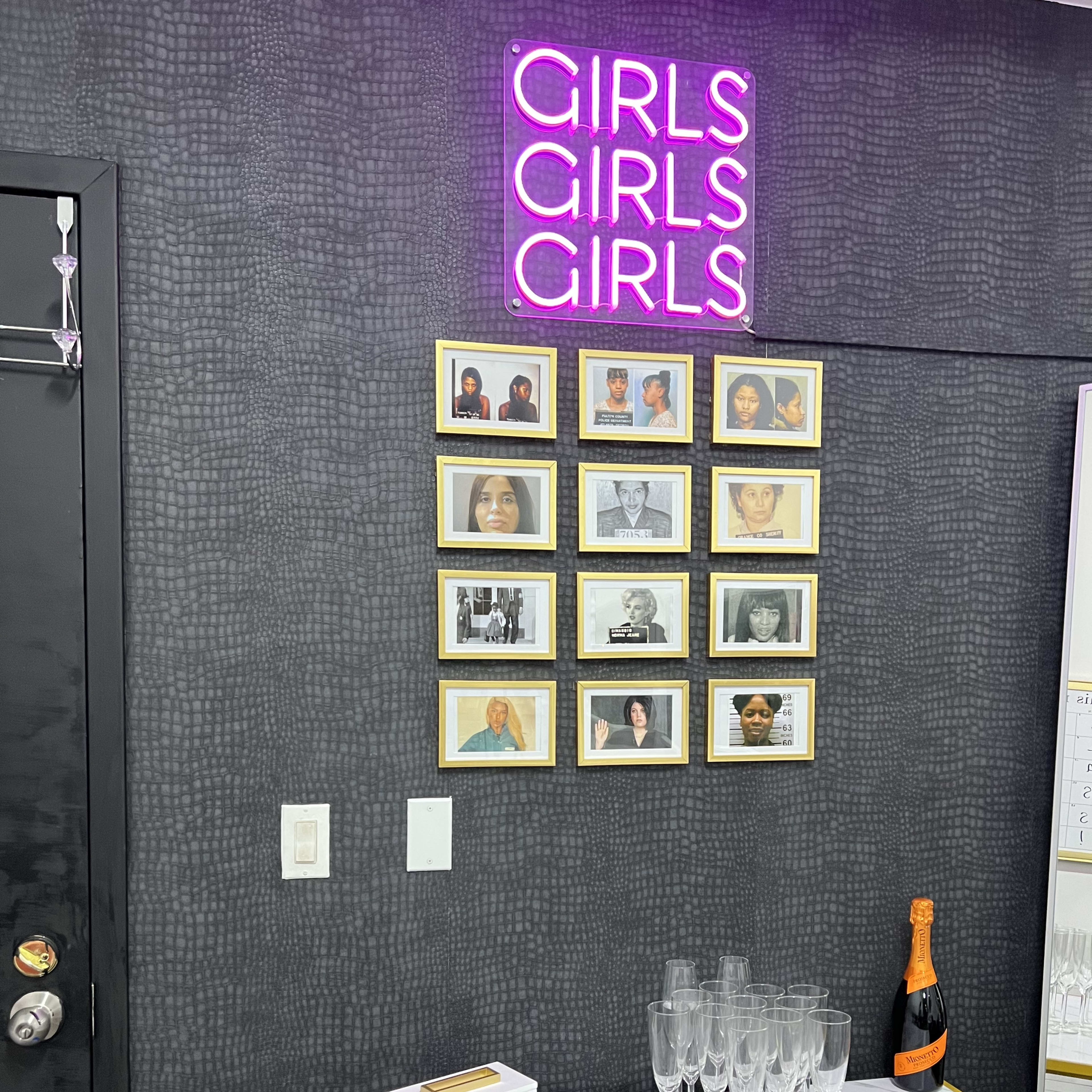 A wall displays a neon sign reading "GIRLS GIRLS GIRLS" above a collection of framed photographs, a table with champagne flutes, and a bottle of champagne.