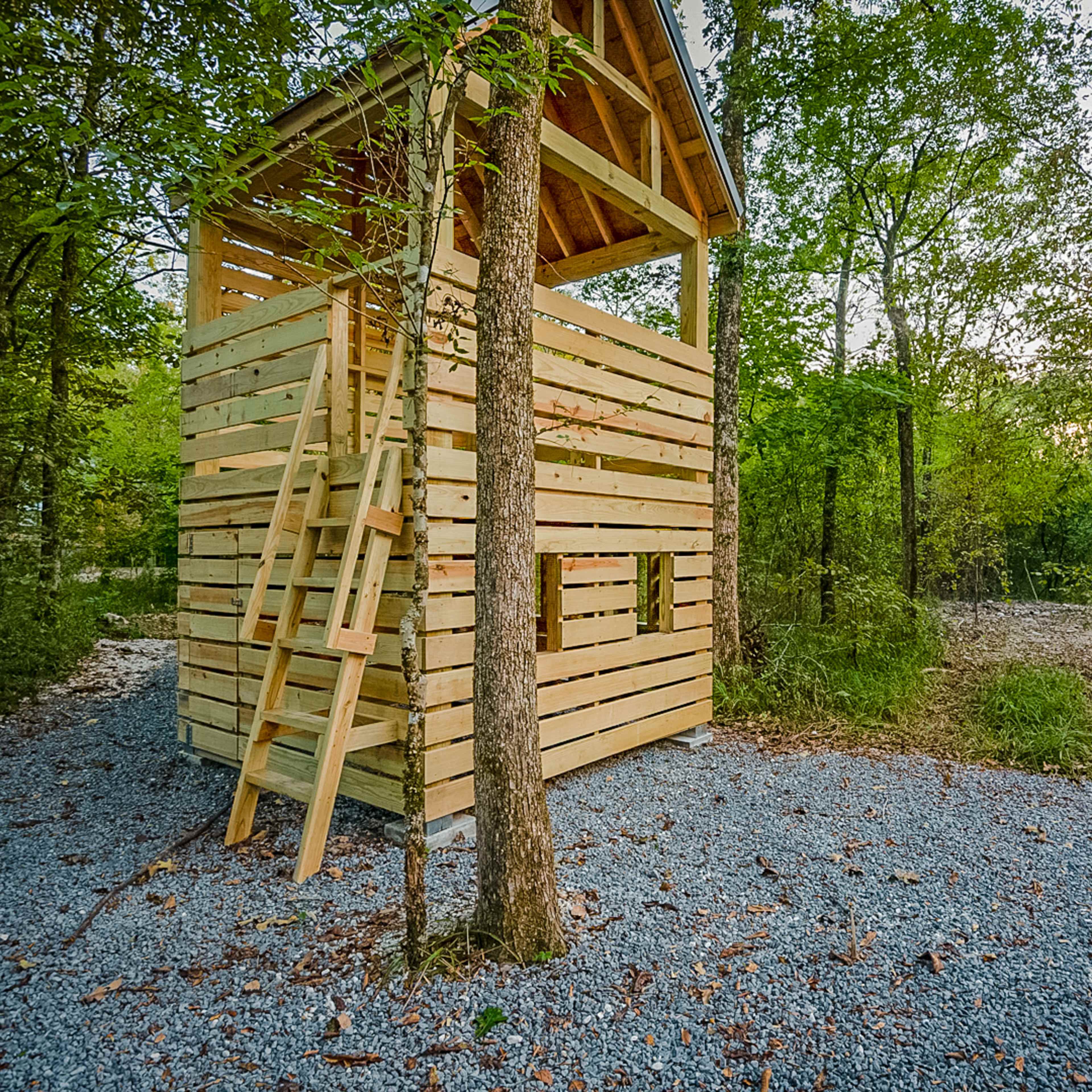 A wooden treehouse with a ladder is situated among trees in a gravel-covered area.