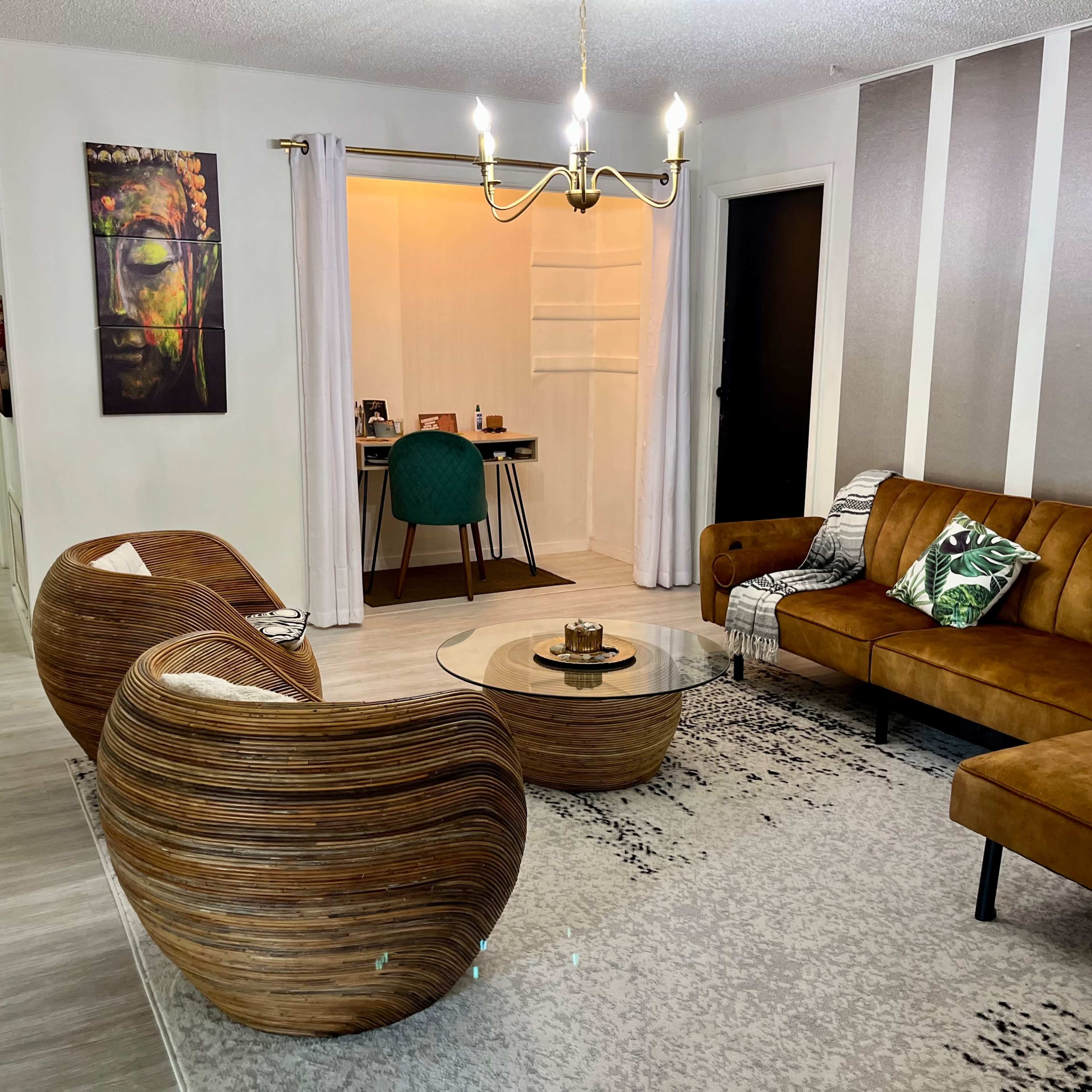 A modern living room features a brown leather sofa, two woven chairs, a glass coffee table, and a workspace area in the background with a green chair.