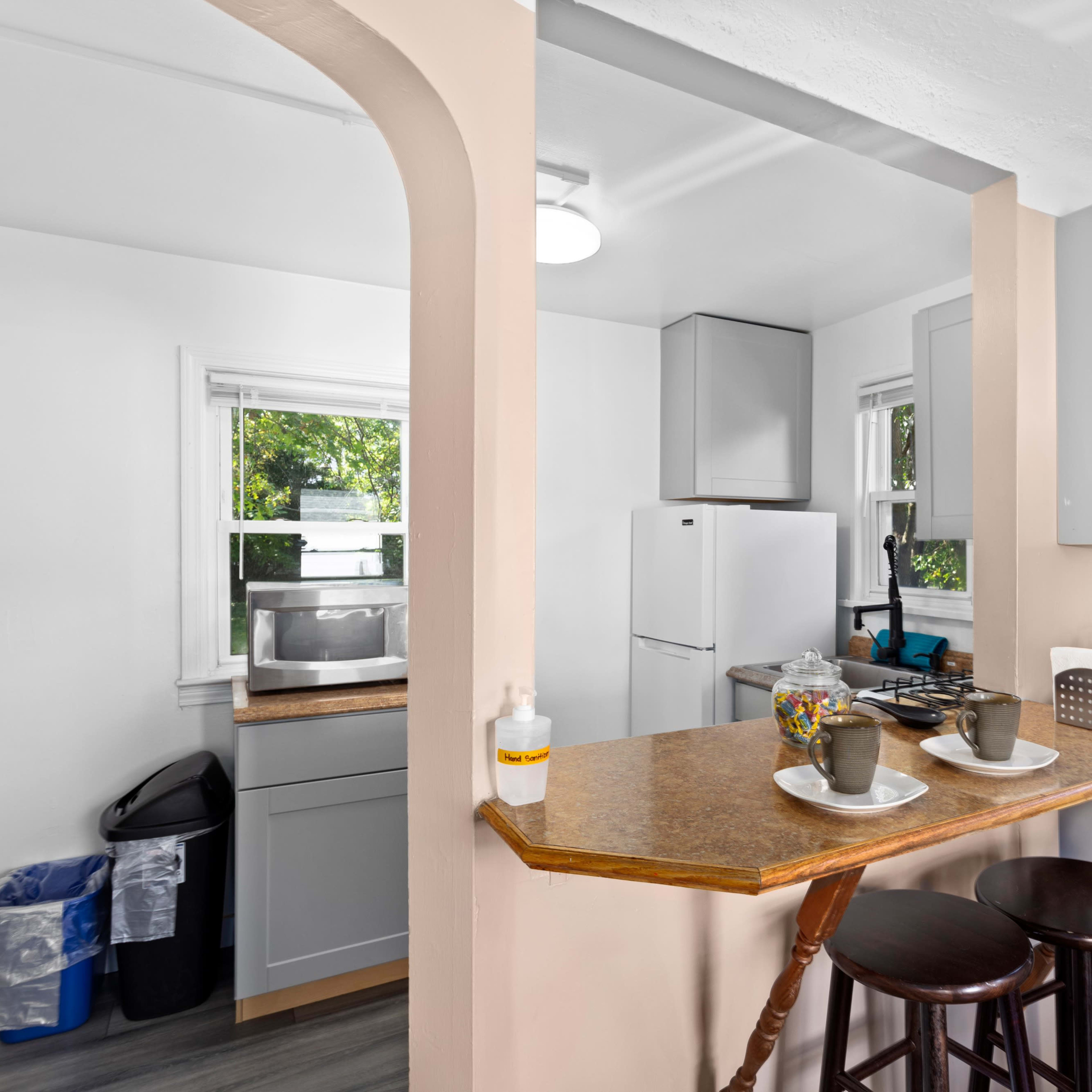 A modern kitchen with light-colored cabinets, a breakfast bar with two stools, and a small dining area visible through an archway.