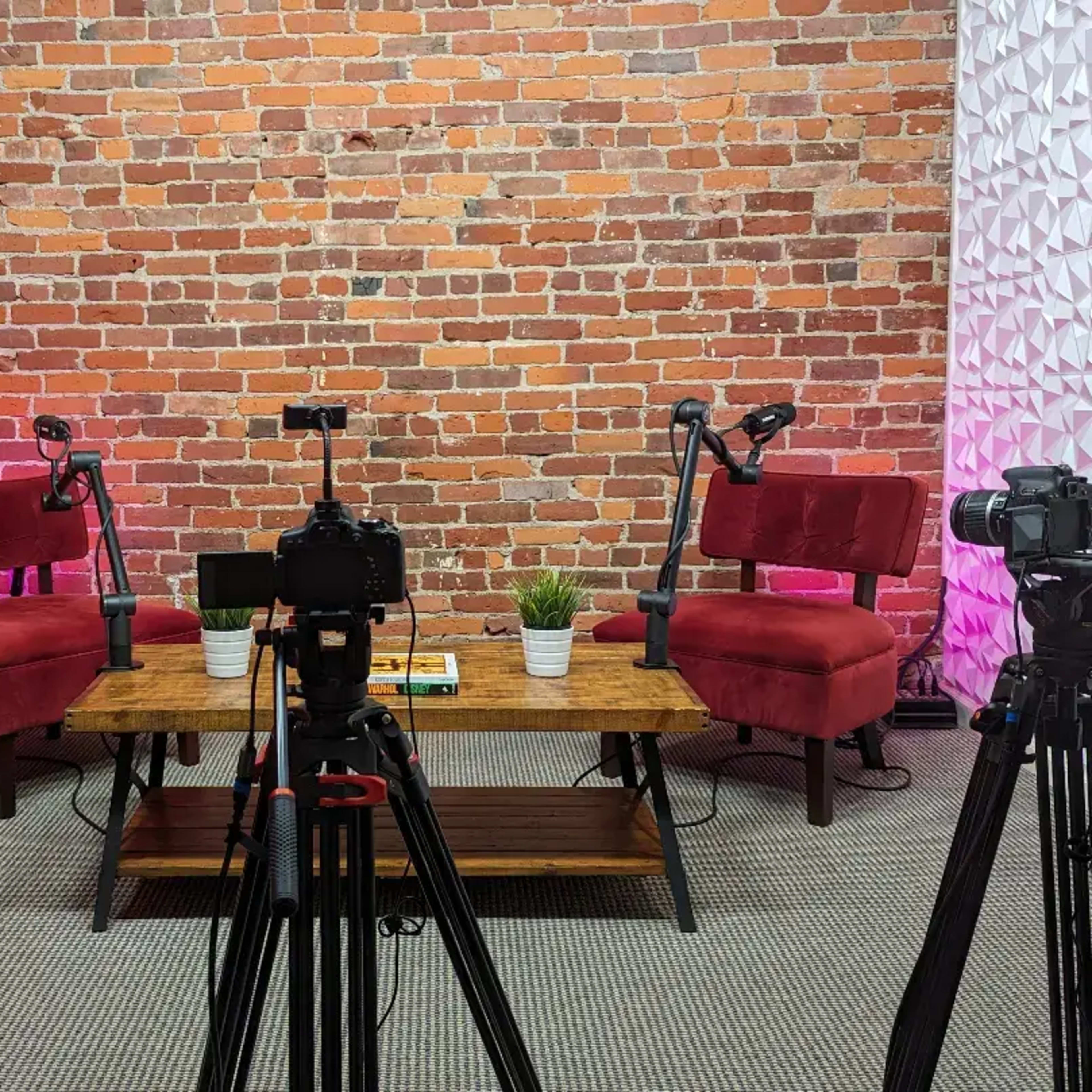 A video recording setup featuring two red chairs and a wooden table, surrounded by three tripods holding cameras, against a brick wall backdrop.