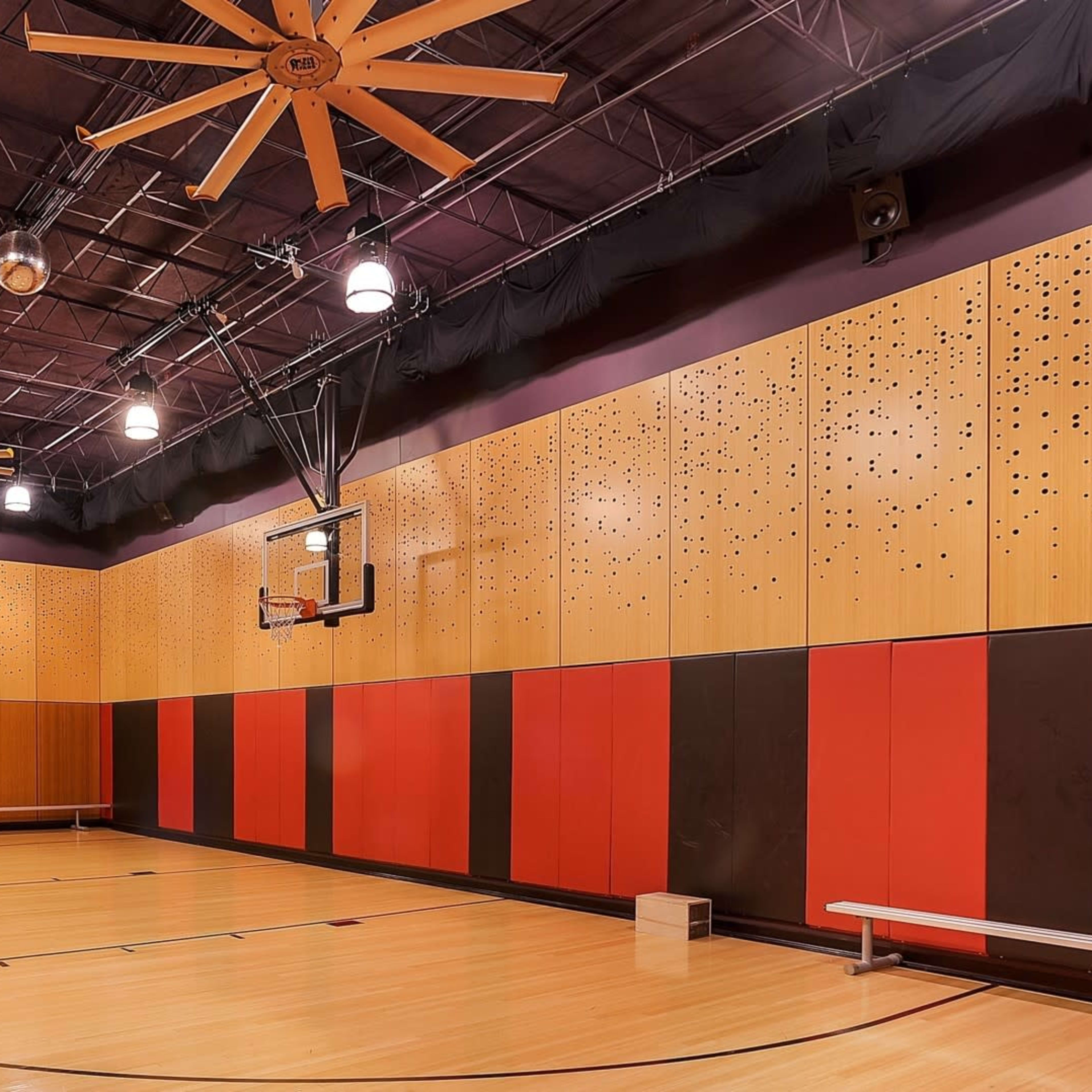 An indoor basketball court with wooden flooring, large ceiling fans, and walls decorated in a pattern of orange, black, and wood paneling.