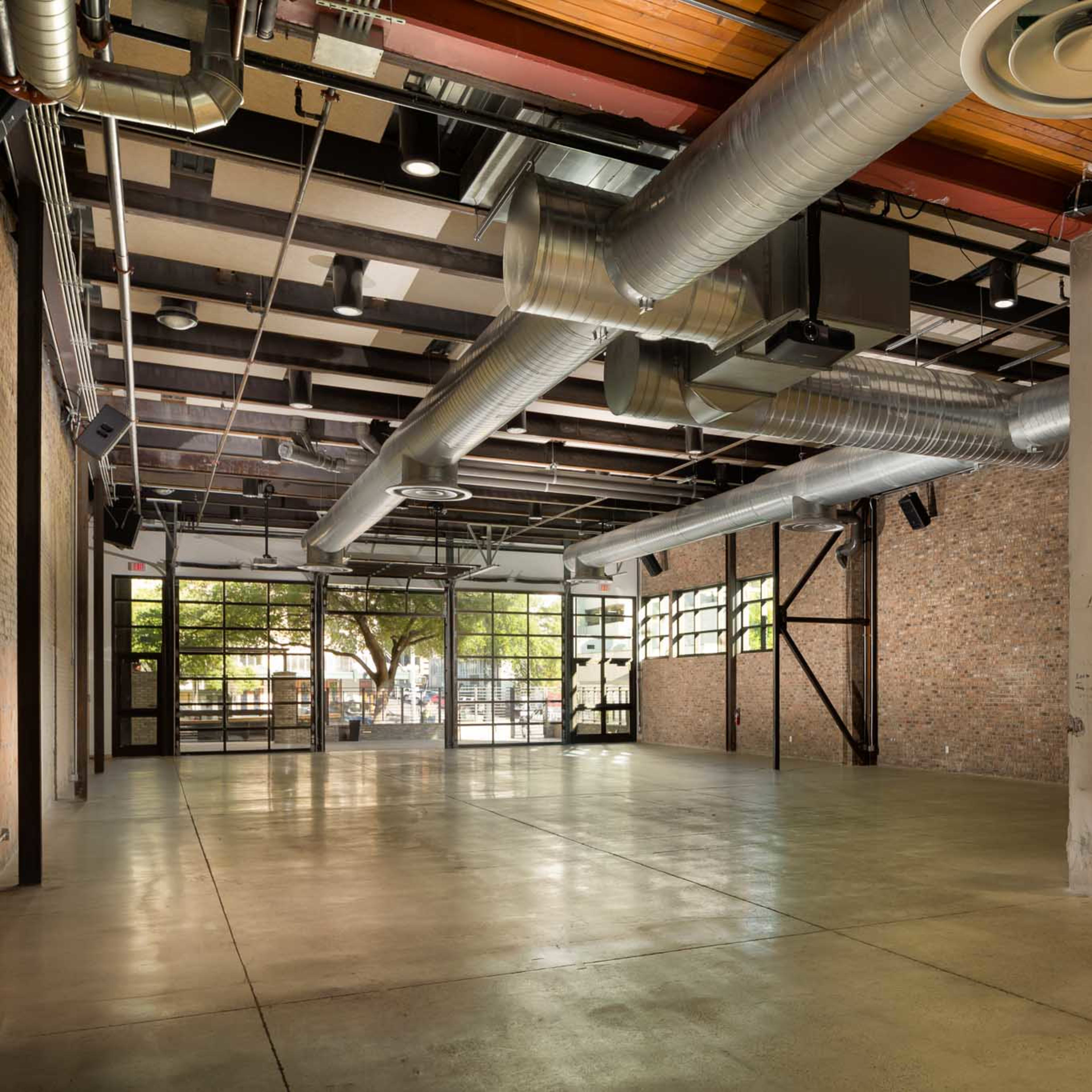 A spacious, industrial-style venue features exposed ductwork, large windows, and a polished concrete floor.