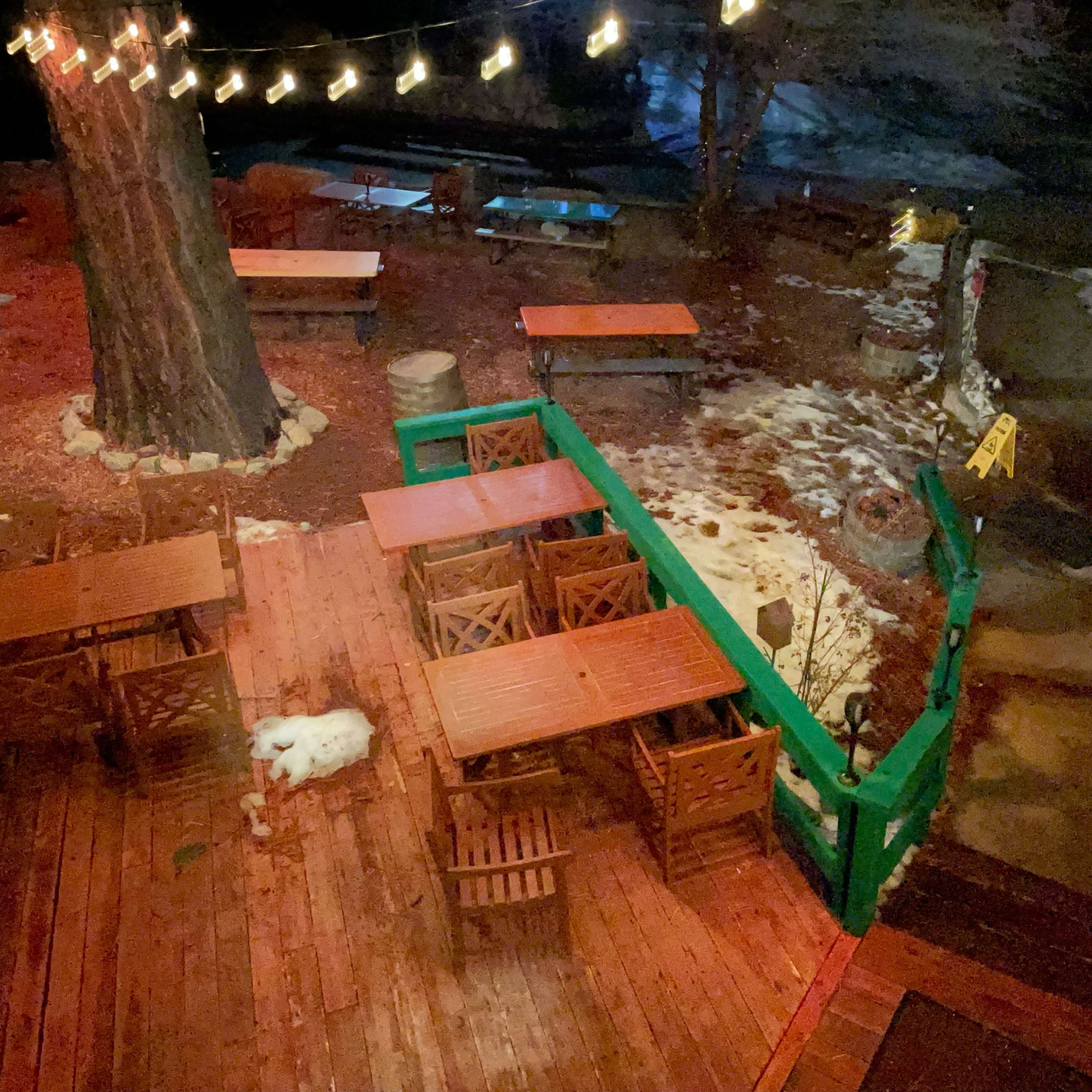 A wooden deck with several tables and chairs is illuminated by string lights, surrounded by a snowy landscape at night.