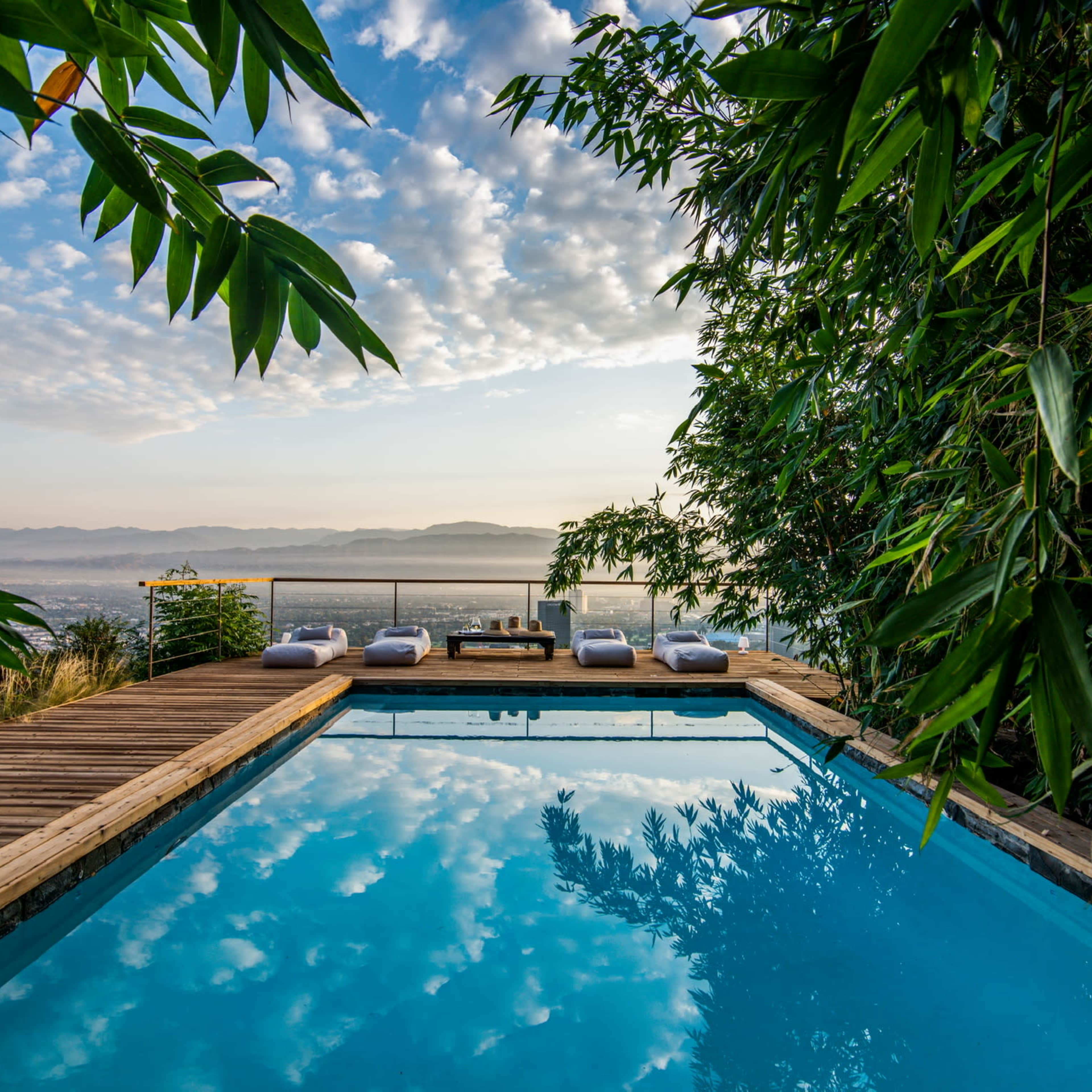 A swimming pool with a wooden deck is surrounded by greenery and offers a view of the mountains under a cloudy sky.
