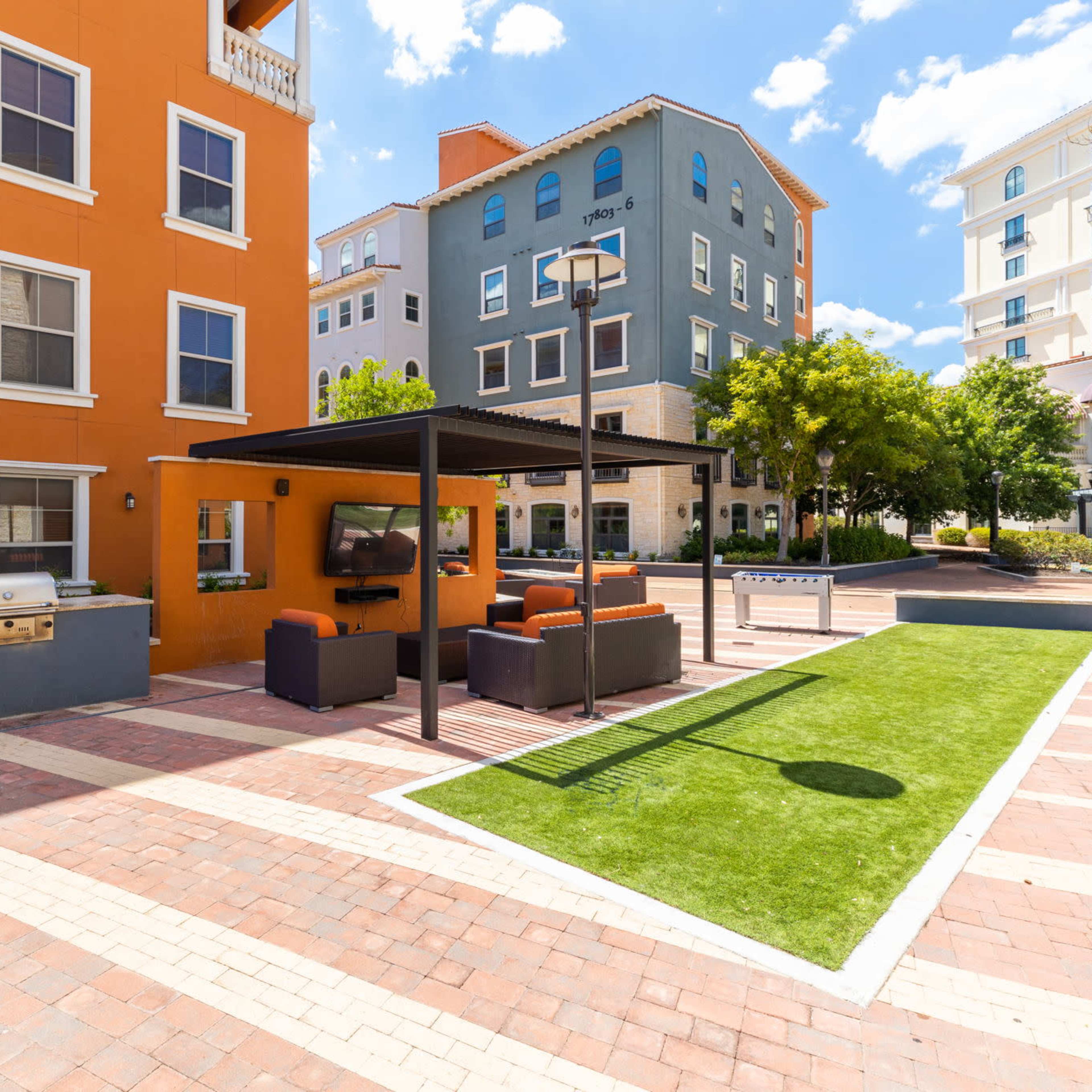 An outdoor communal area with seating, a covered grill station, and landscaped green space surrounded by colorful apartment buildings.