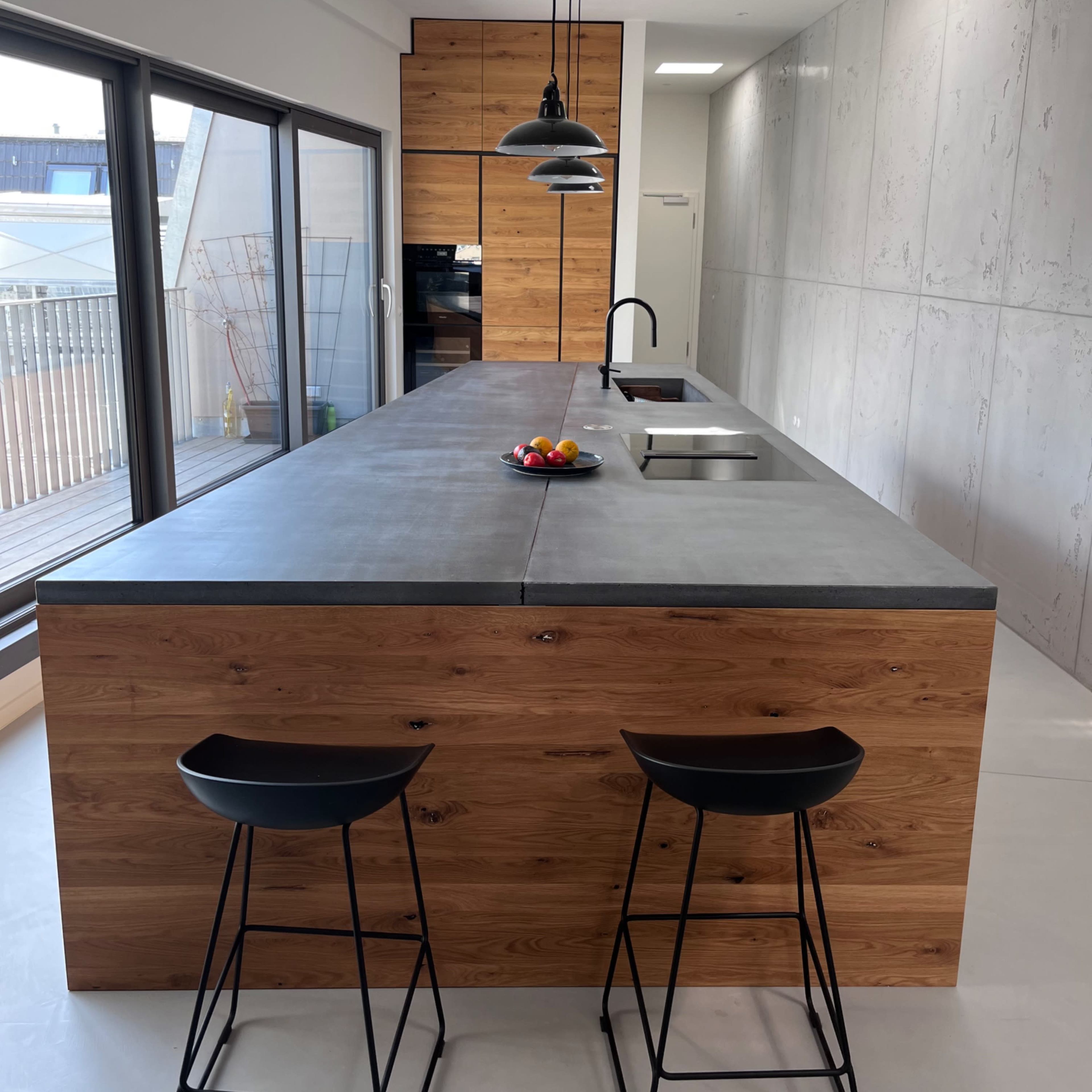 A modern kitchen with a large concrete countertop, wooden cabinetry, and two black stools.