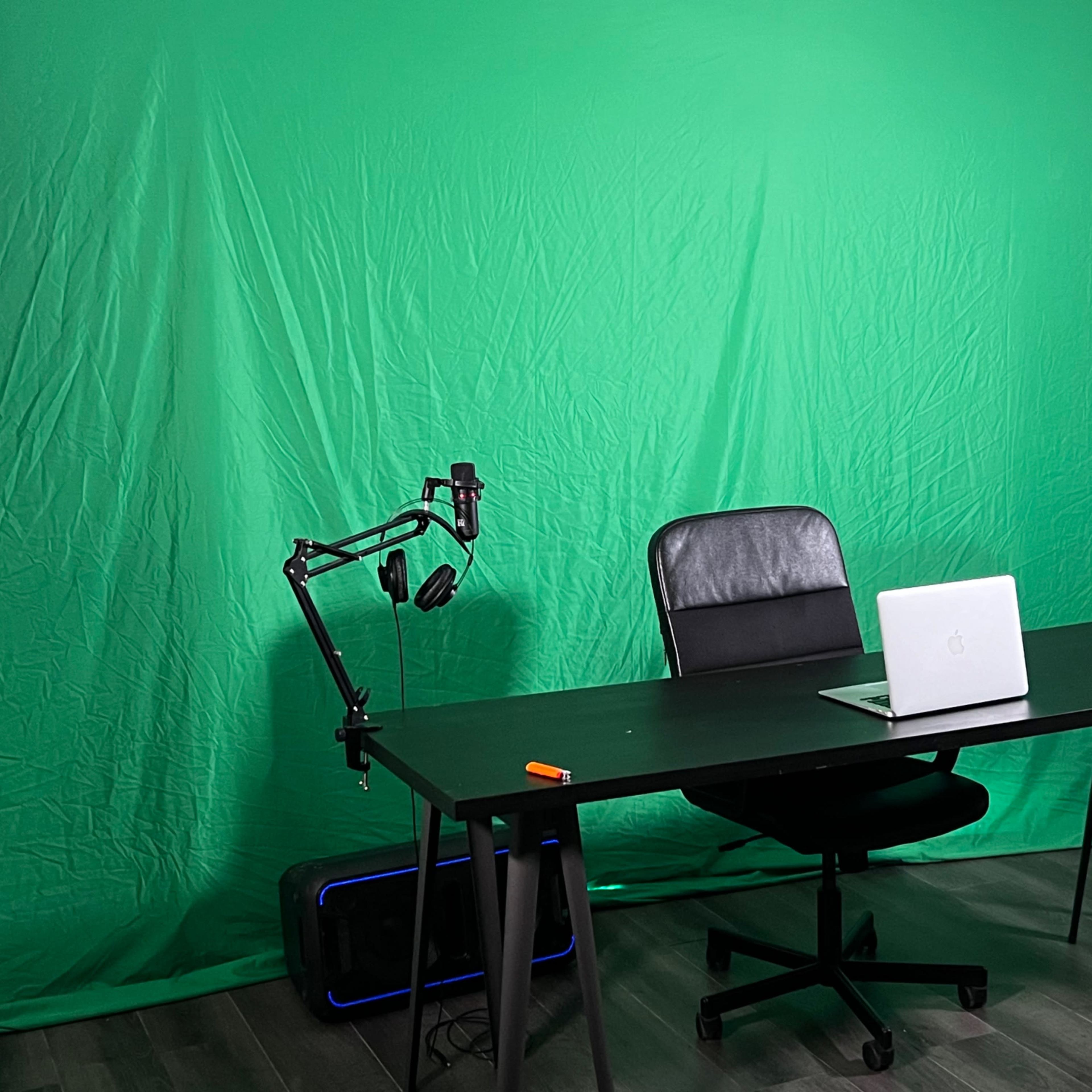 A desk with a laptop and a microphone arm set up in front of a green backdrop.