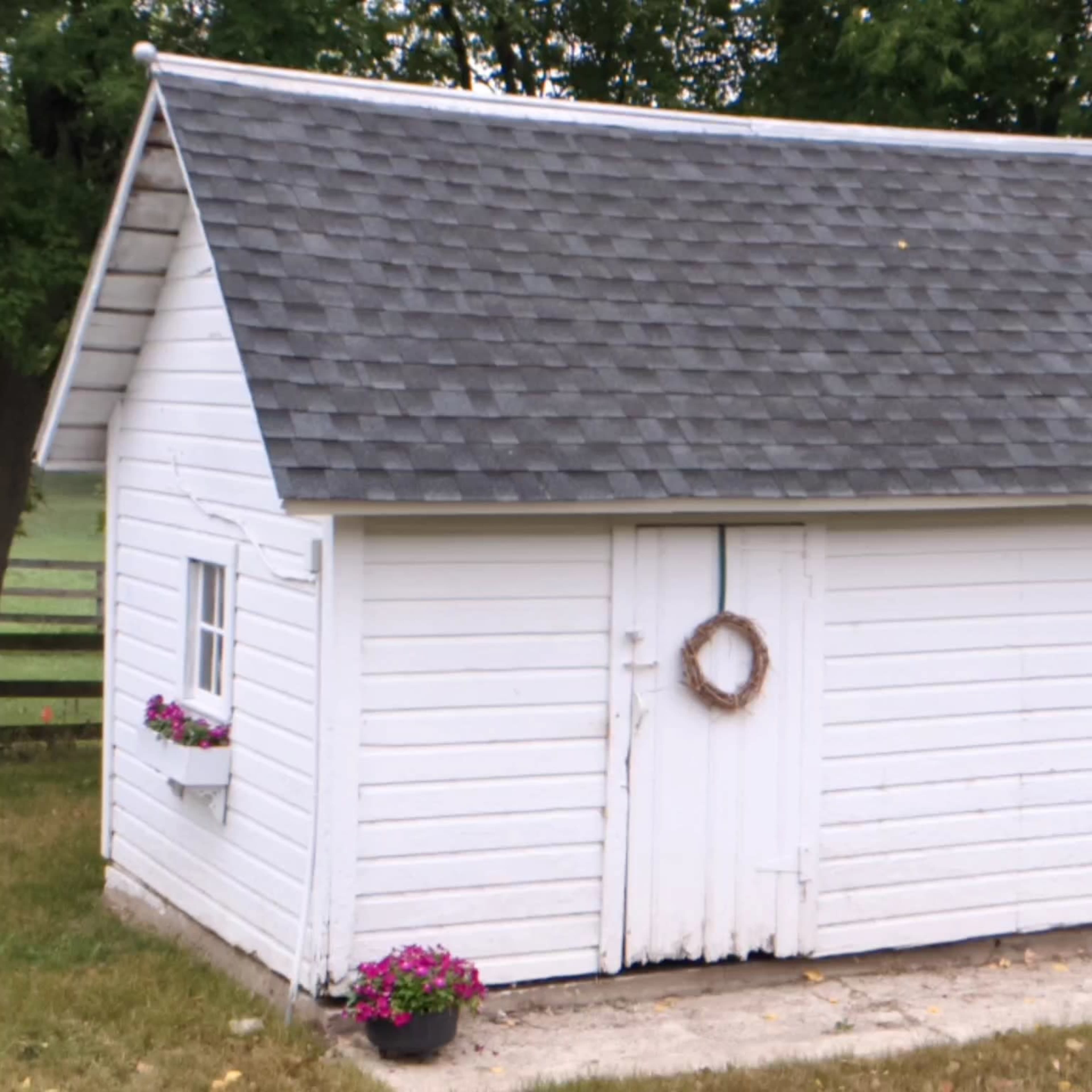 A small white shed with a gray roof, featuring a wreath on the double doors and flower pots at its base.