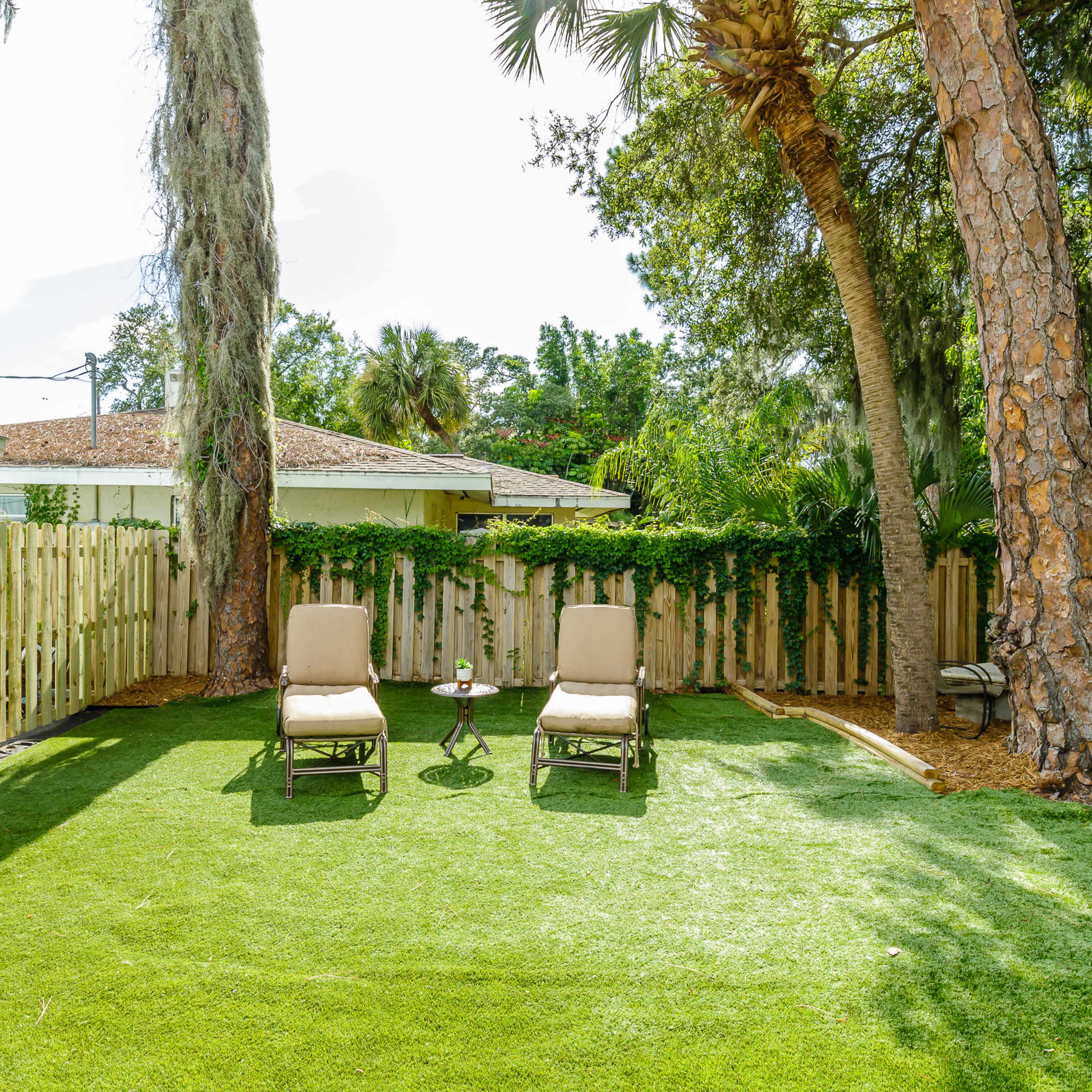 A backyard with two lounge chairs and a small table on artificial grass, surrounded by trees and a wooden fence.