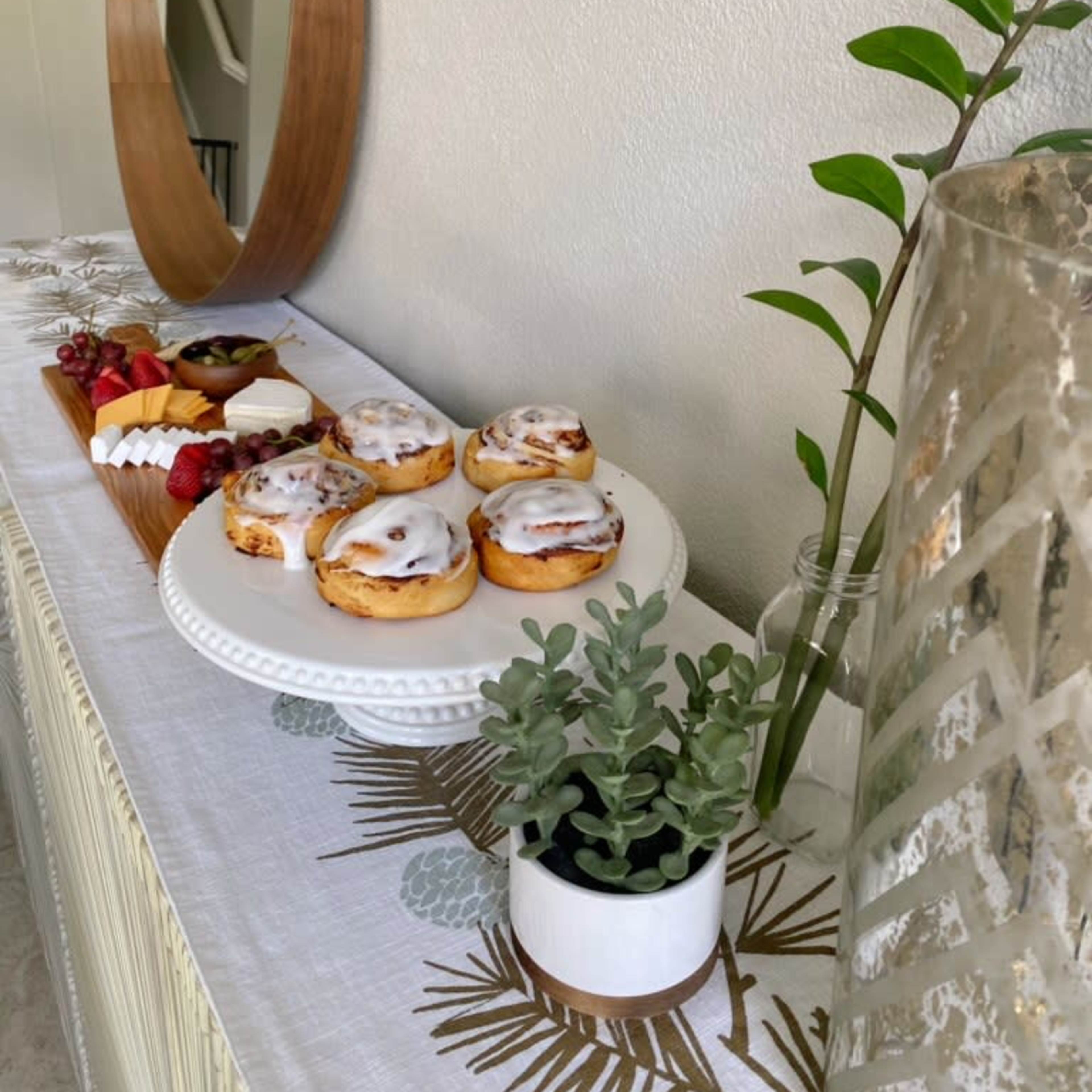A table is set with a mirror, a small plant, a decorative vase, and a plate of cinnamon rolls alongside fruits and cheese on a wooden tray.