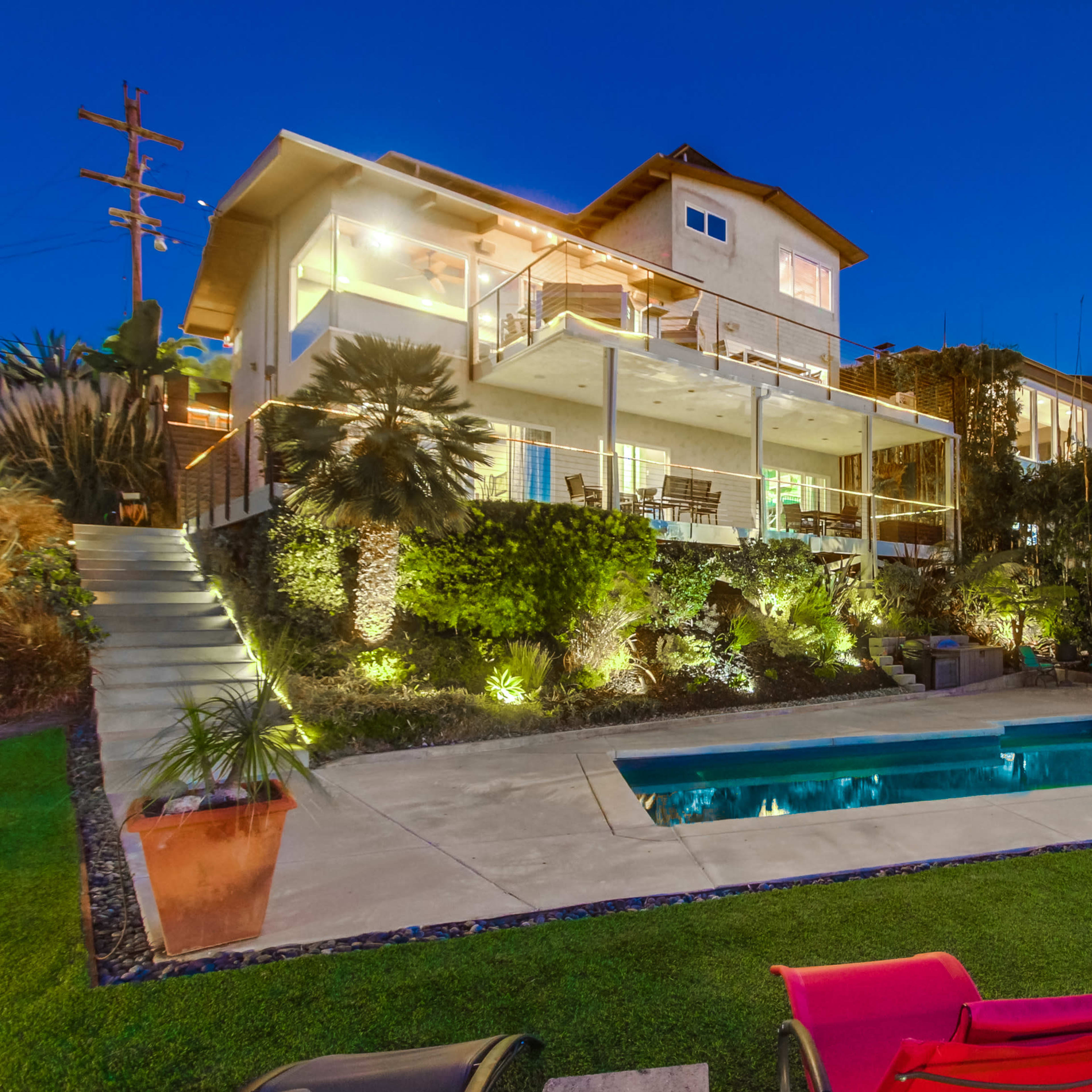 A two-story house with multiple balconies overlooks a swimming pool surrounded by landscaped greenery in the evening.