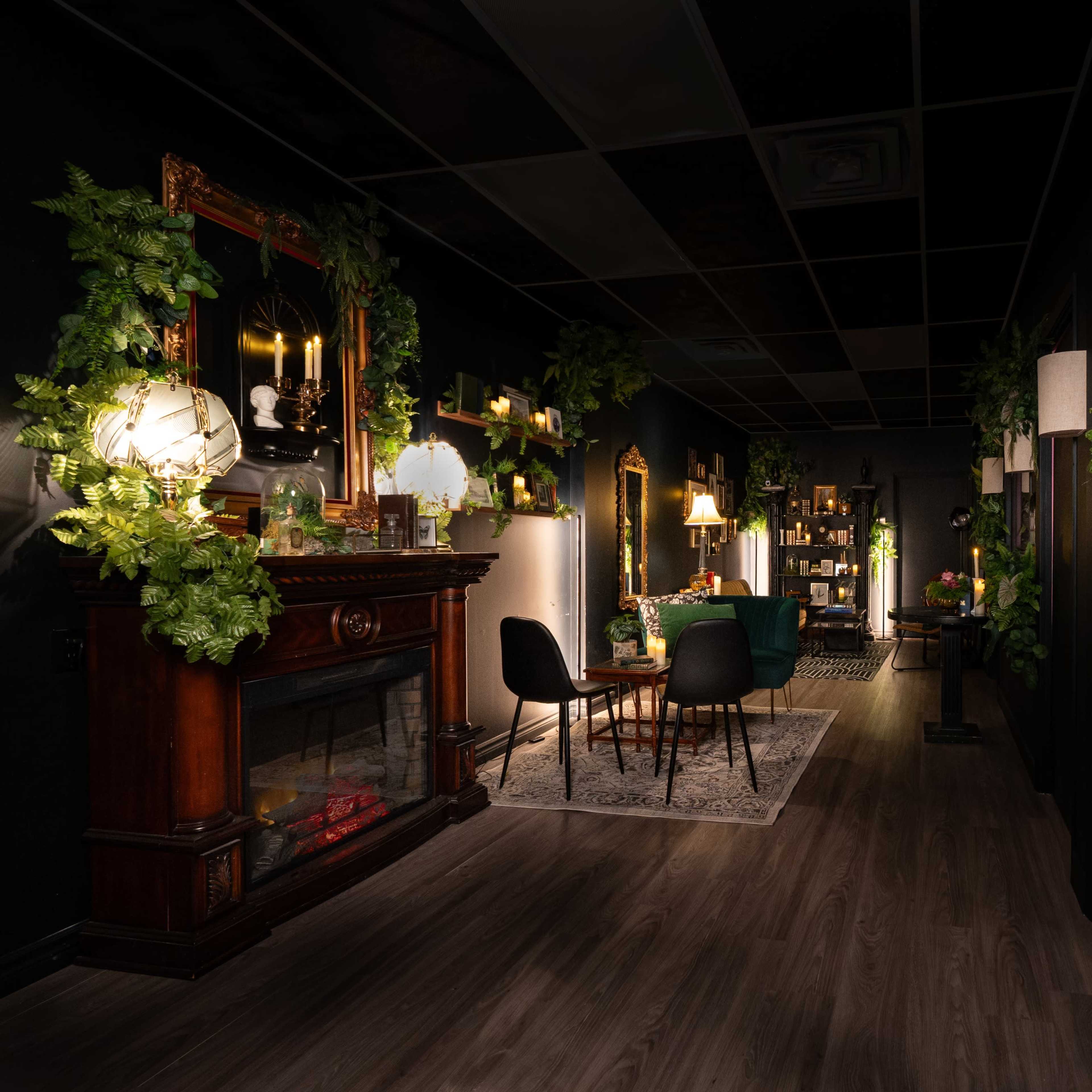 A dimly lit room with dark walls, featuring a fireplace, mirrors, various plants, and elegant furniture arranged in a cozy setting.