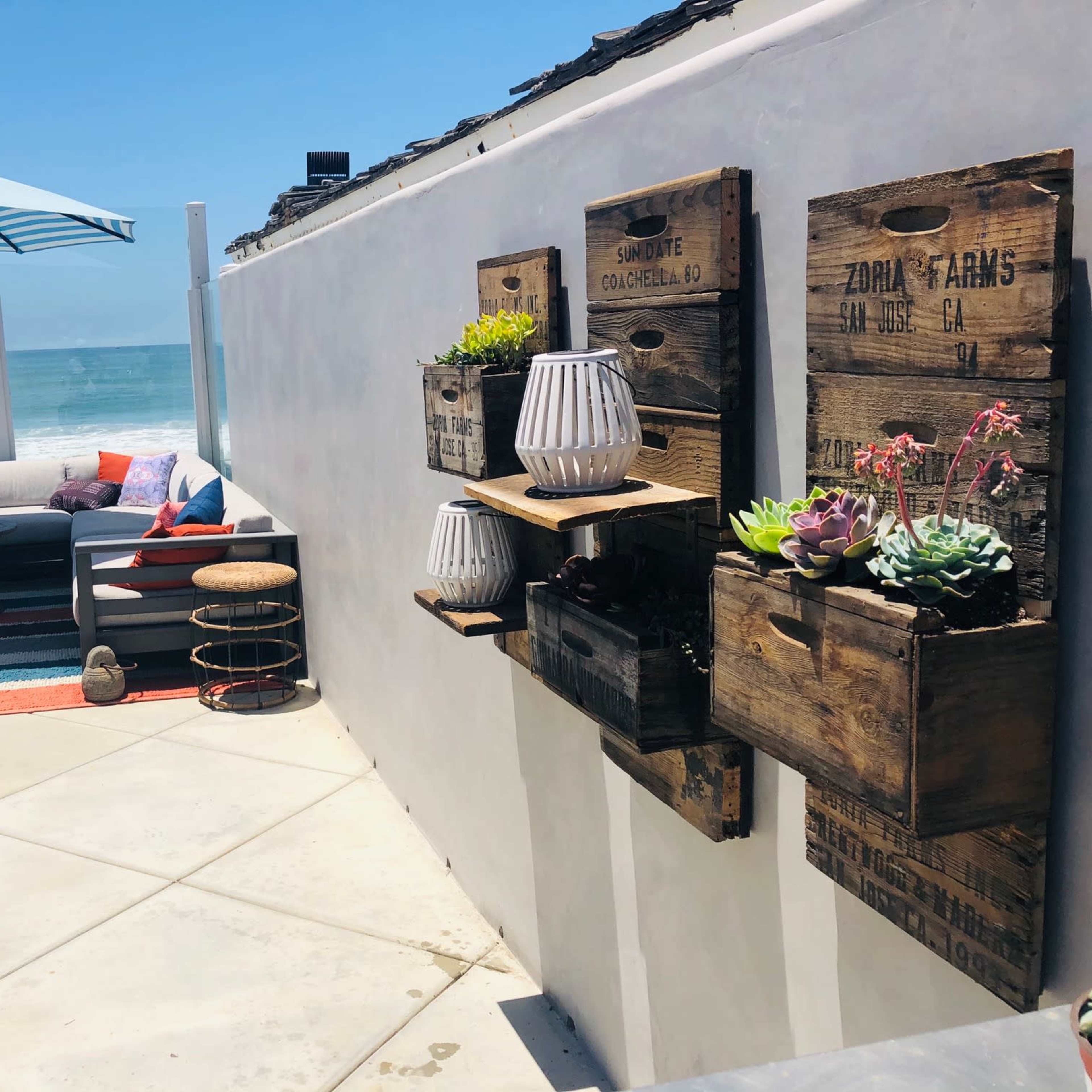 A coastal patio area with a seating arrangement under a blue umbrella, flanked by wall-mounted planters filled with plants, and a view of the ocean in the background.
