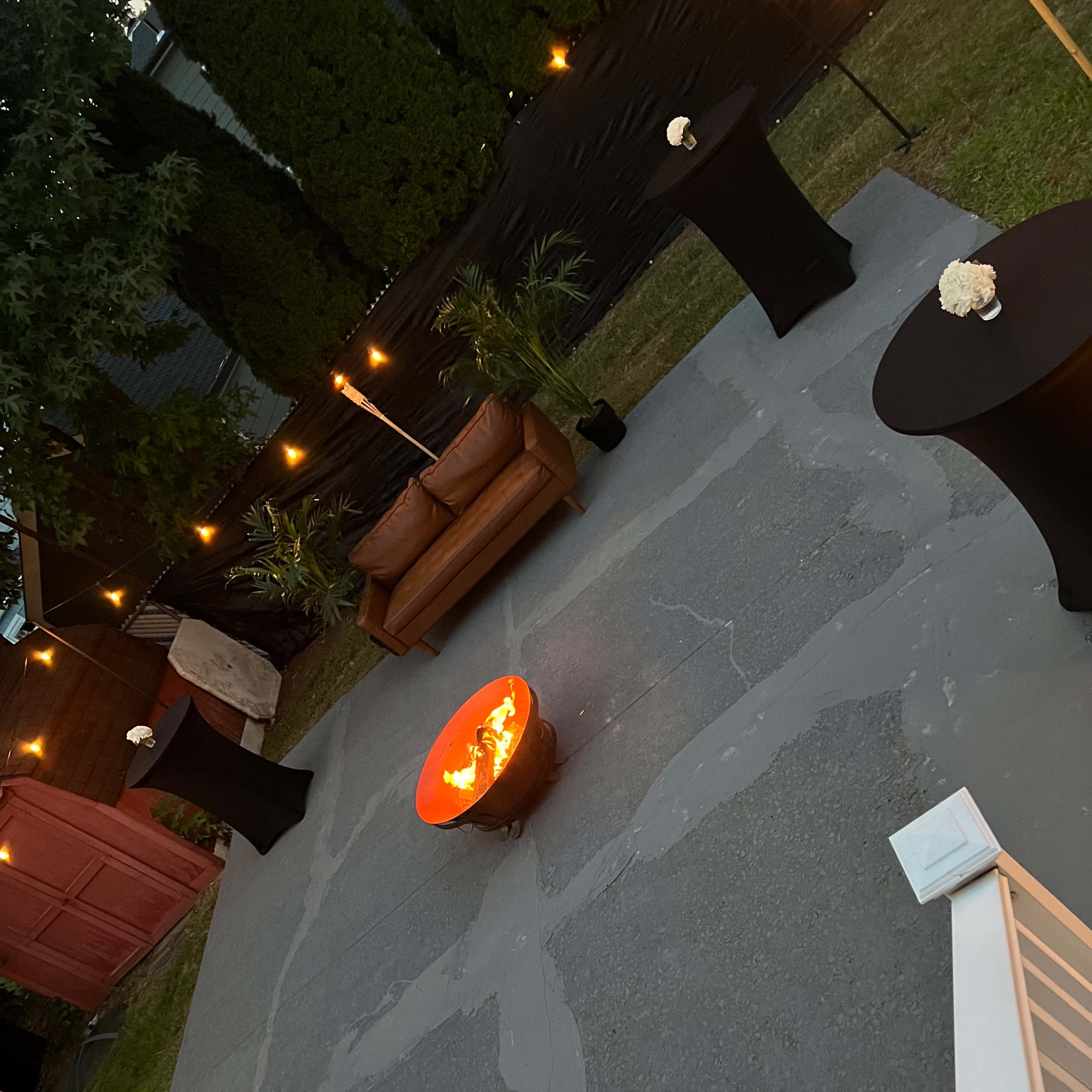 An outdoor patio area with a circular fire pit at the center, surrounded by black cocktail tables and a brown couch, illuminated by string lights.