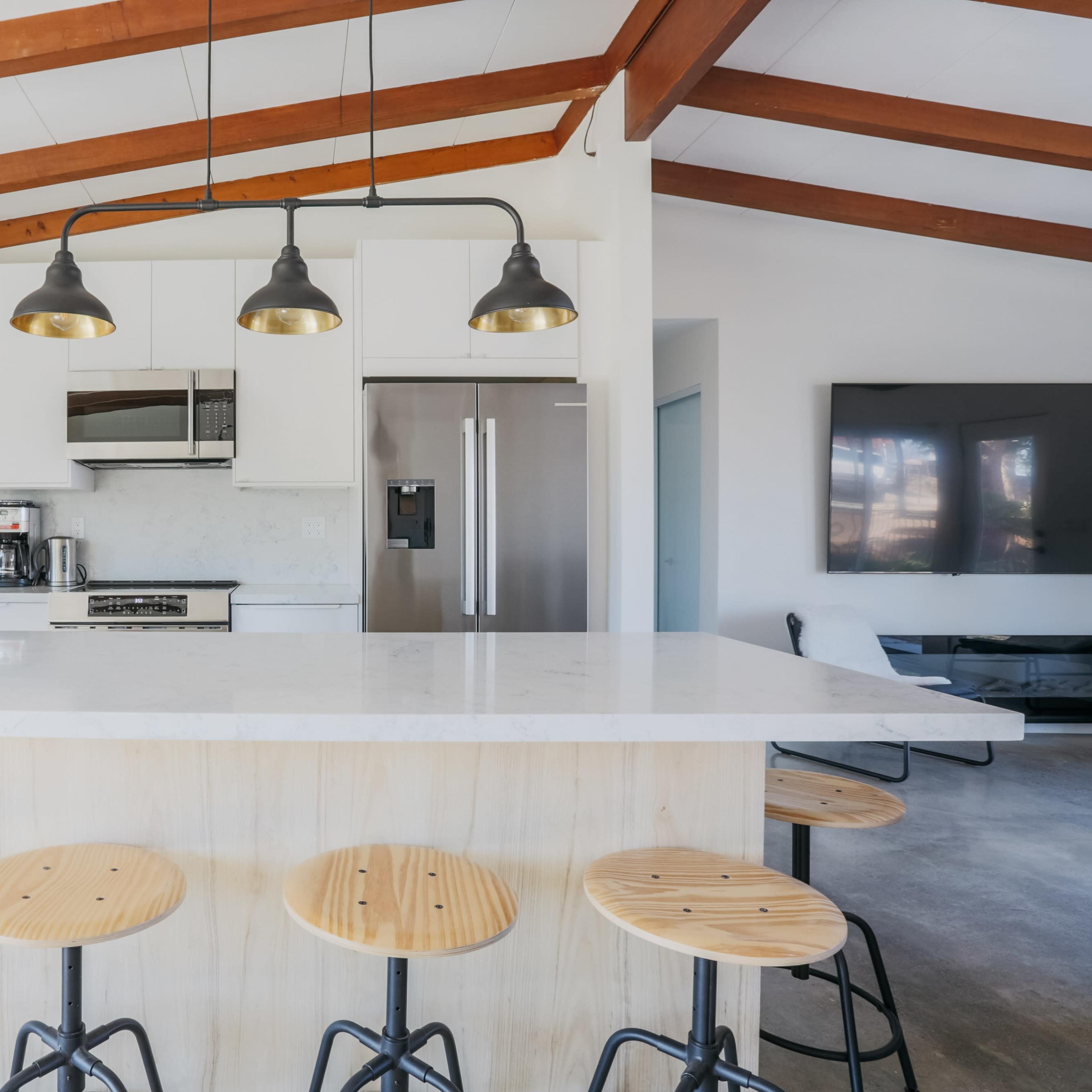 A modern kitchen features a large island with wooden stools and overhead pendant lights, set against white cabinetry and a stainless steel refrigerator.