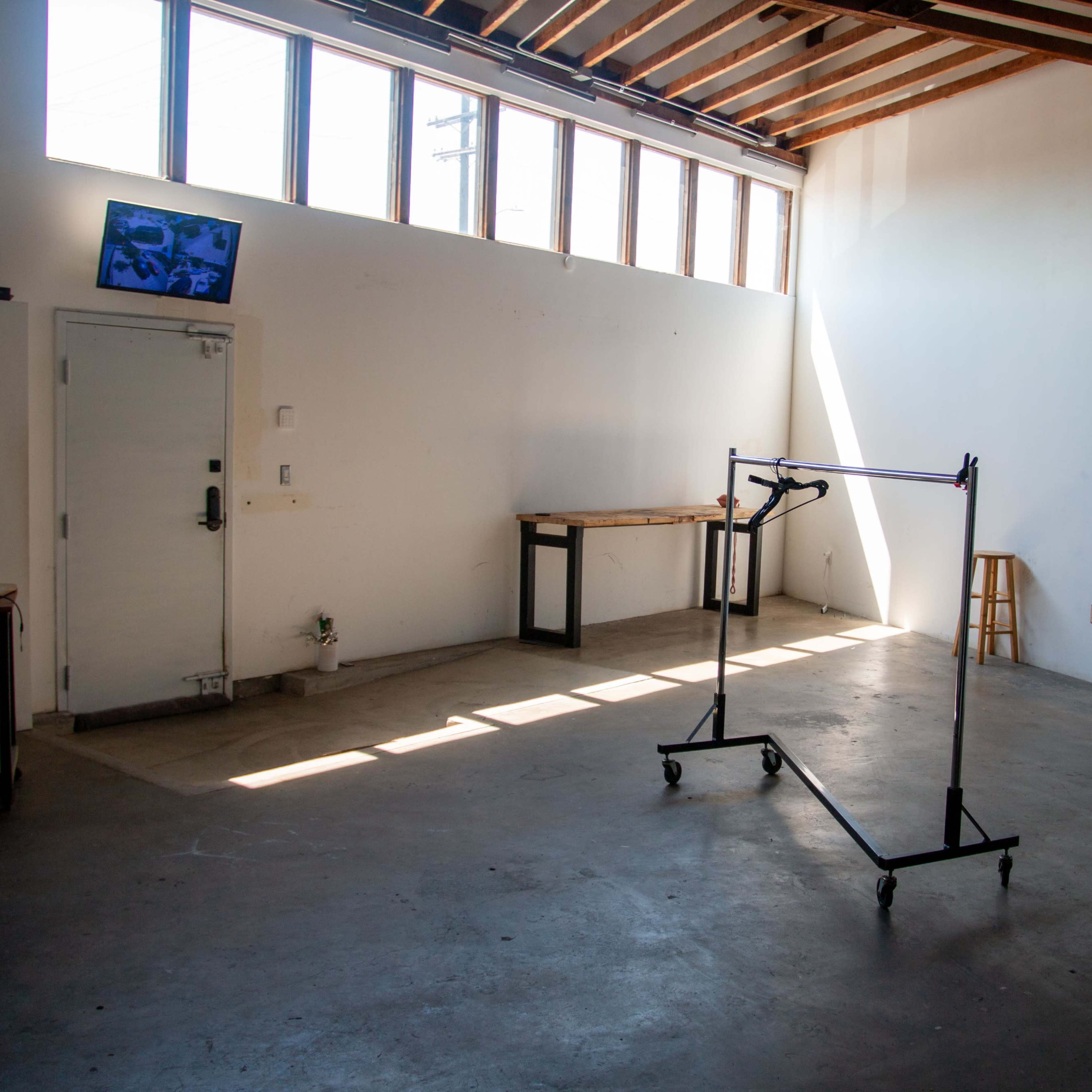 An empty room features a clothing rack on wheels, a table, a stool, and a television mounted on the wall, with light streaming in through large windows.