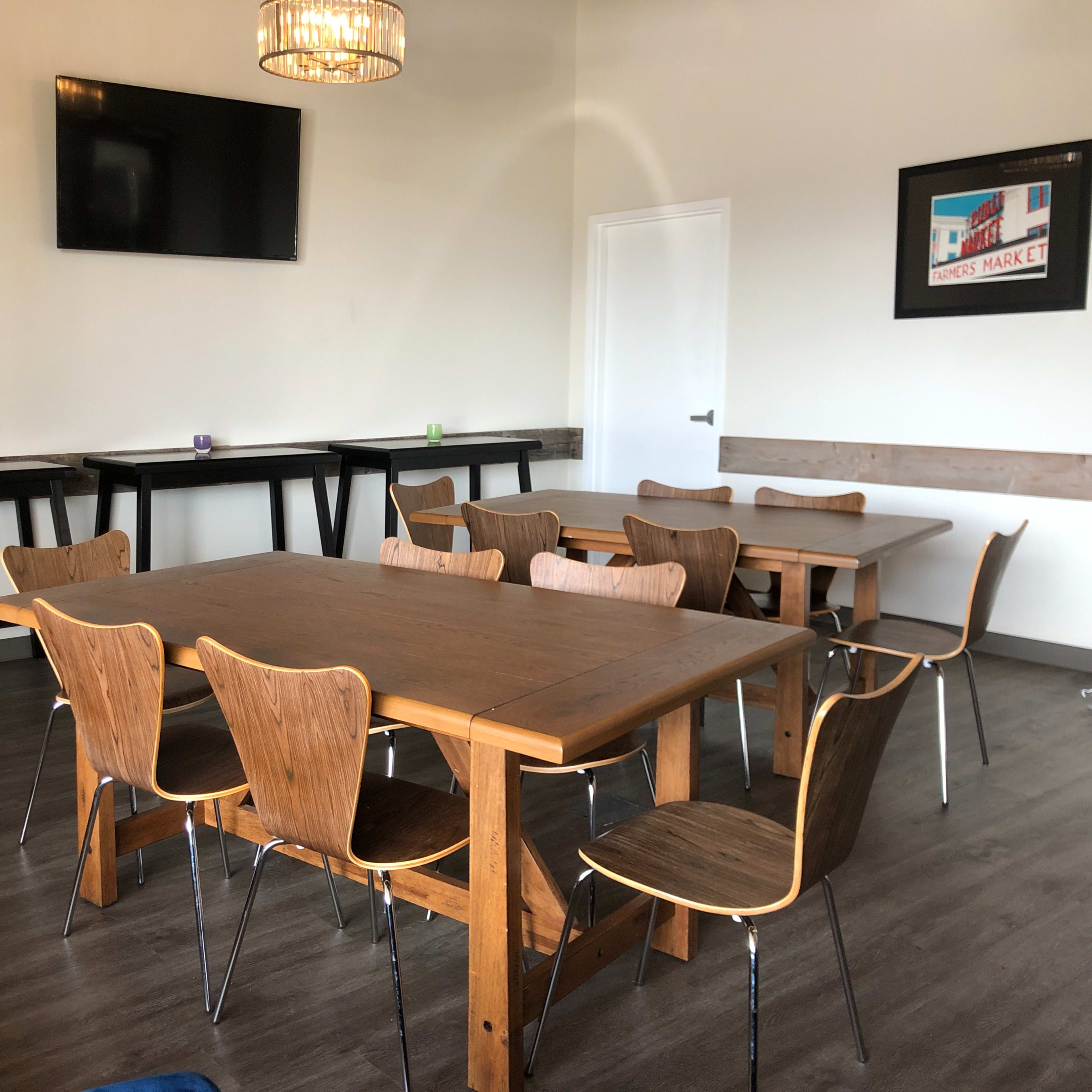 A minimalistic meeting room with several wooden tables and chairs arranged around the space, a wall-mounted TV, and decorative wall art.