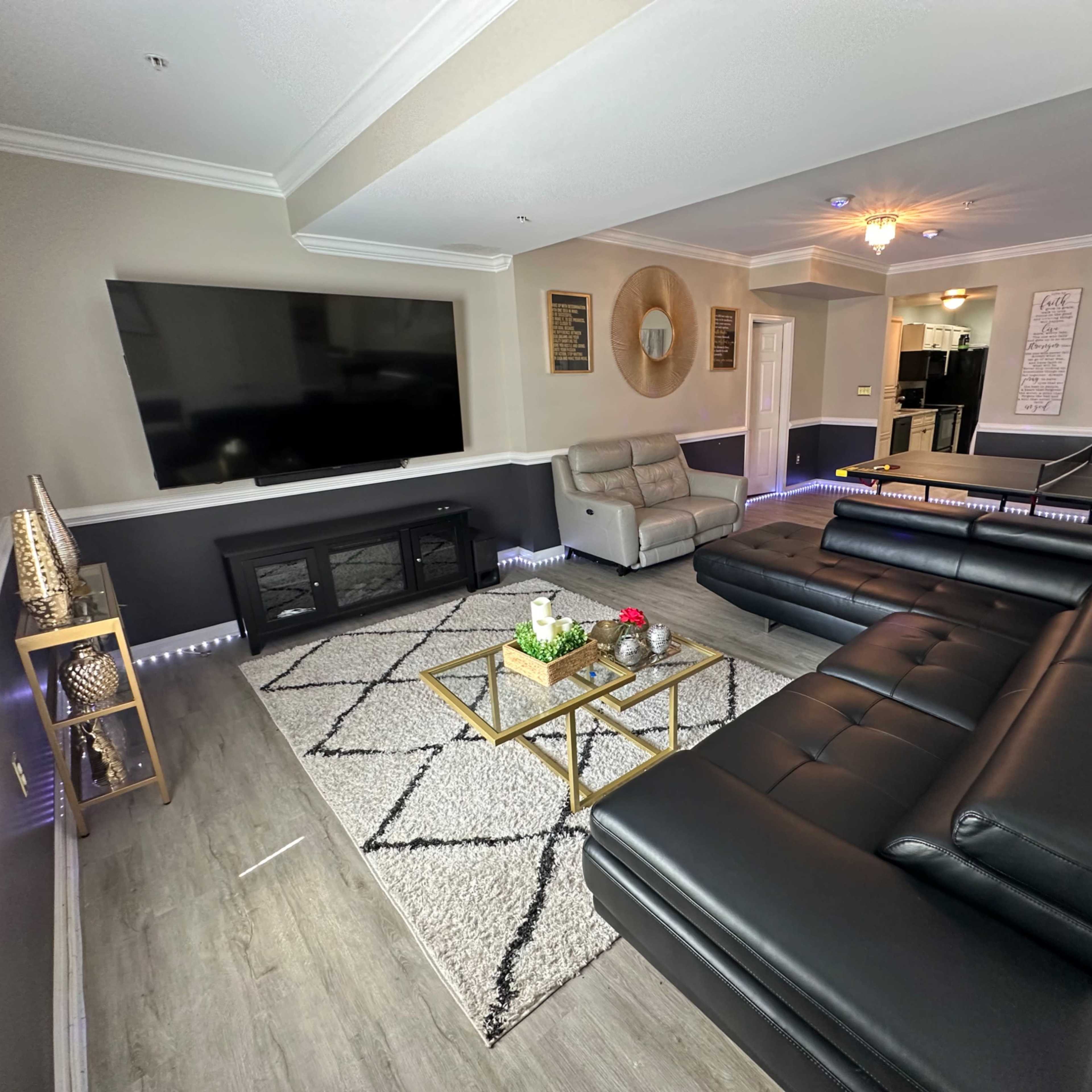 The scene features a modern living room with a large dark sectional sofa, a gold coffee table, a mounted flat-screen TV, and minimalistic decor against light-colored walls.