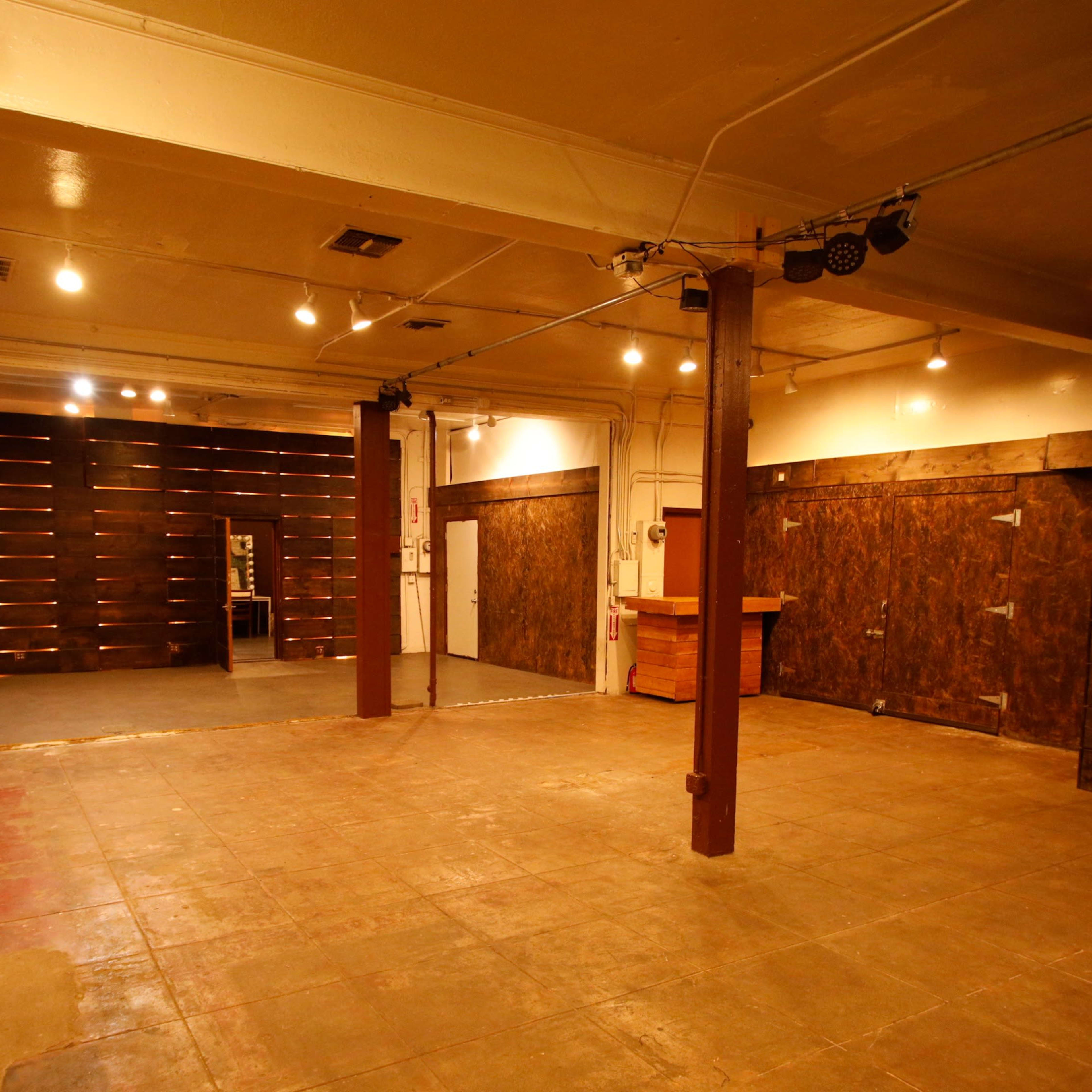 A spacious, empty interior with wooden walls and a concrete floor, featuring overhead lighting and a bar area in the corner.
