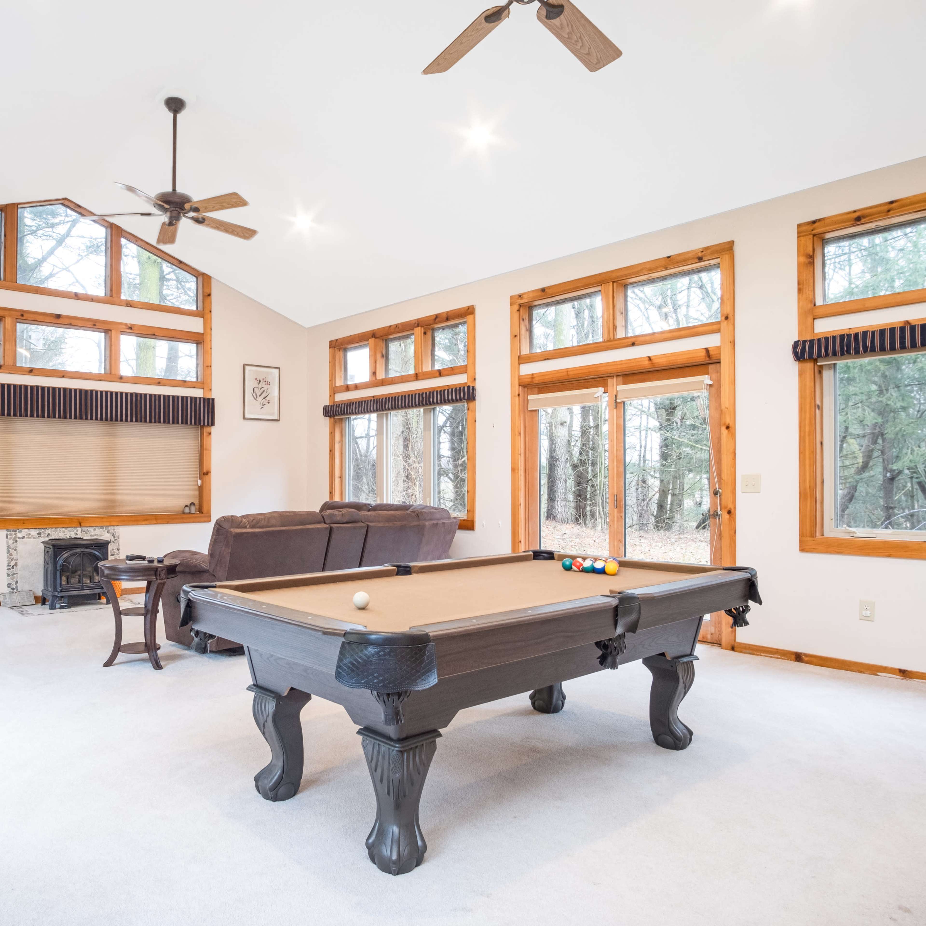 The spacious room features a pool table, large windows, and a flat-screen TV, with views of the surrounding forest.