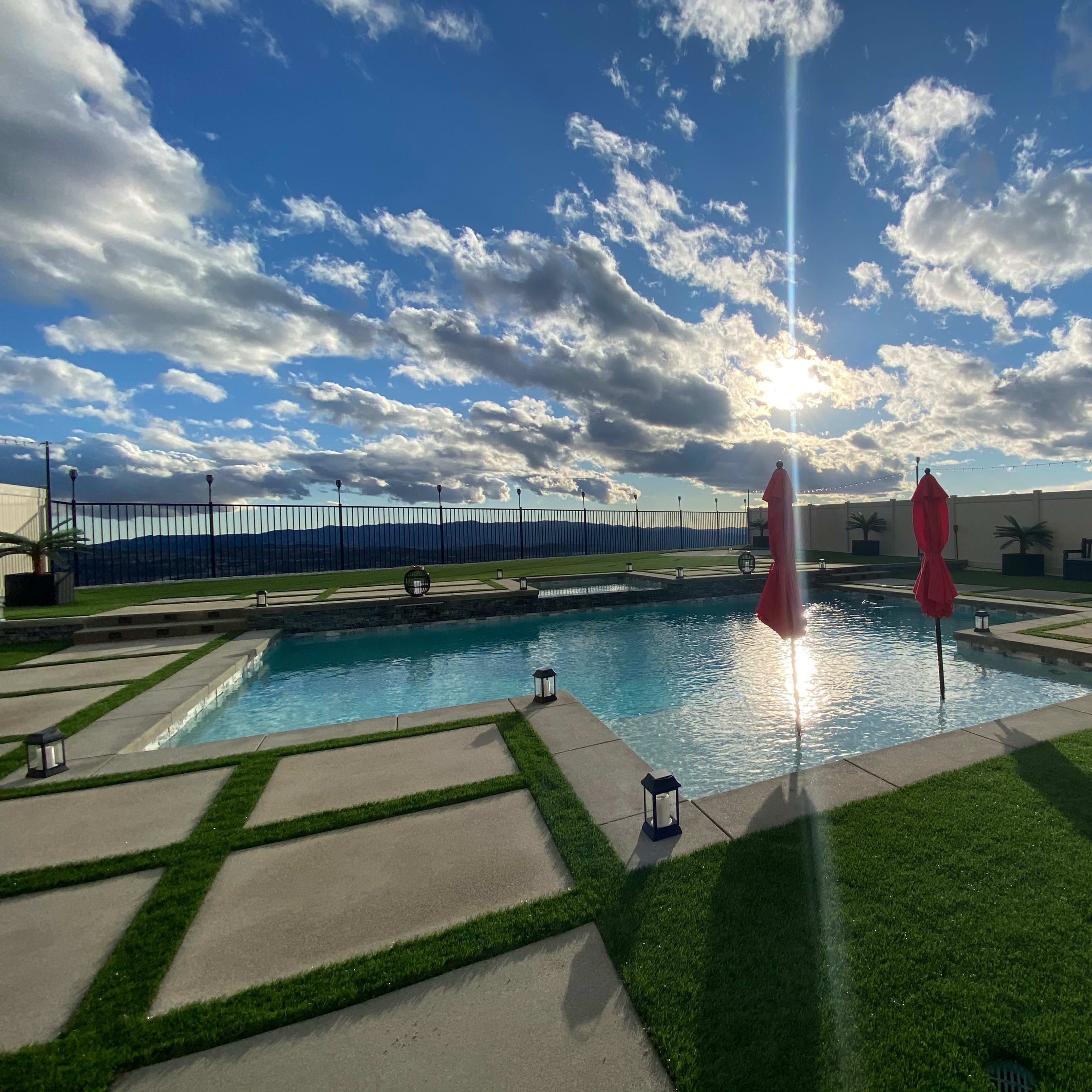 A landscaped pool area with two red umbrellas and a backdrop of cloudy skies at sunset.