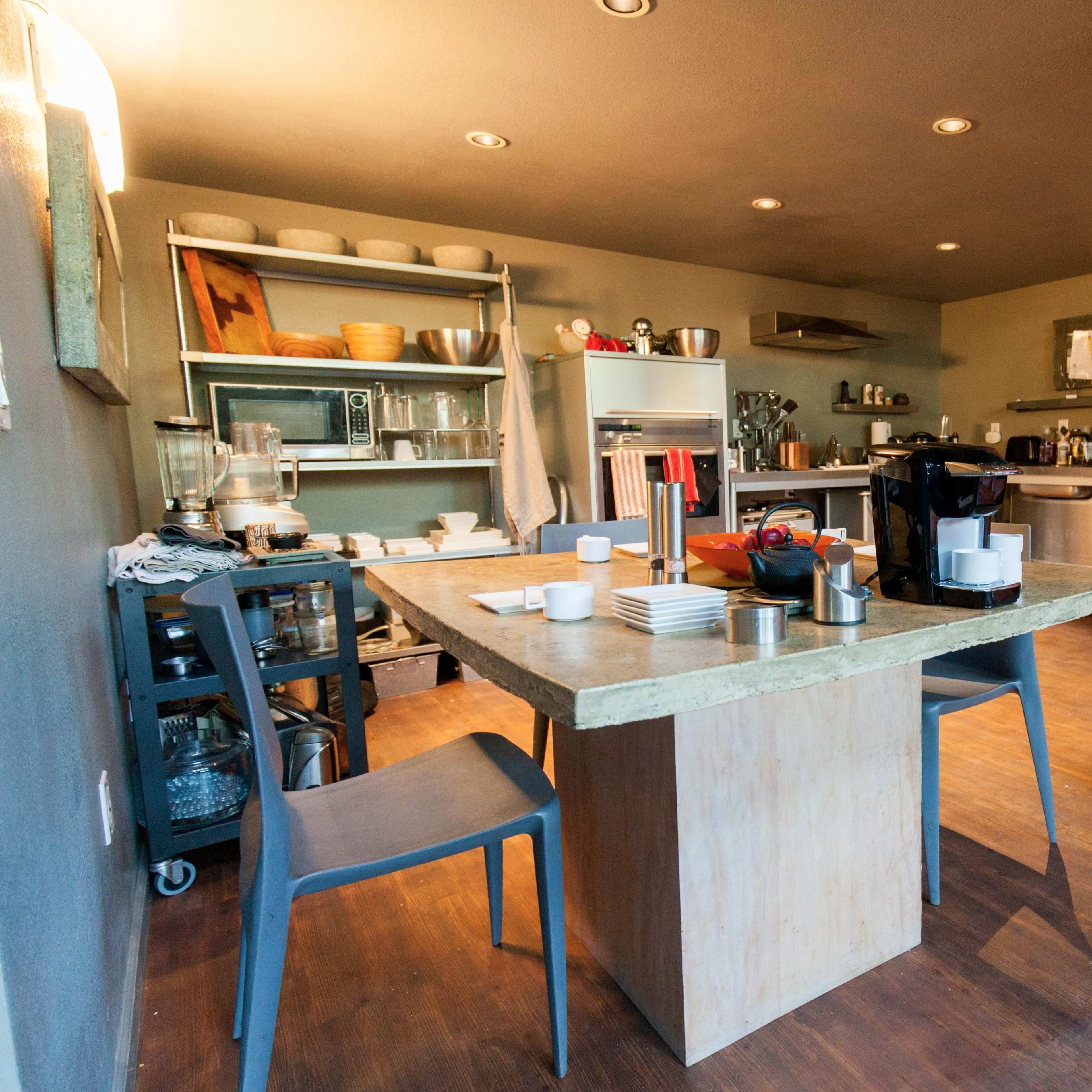 A modern kitchen features a wooden table surrounded by chairs, with stainless steel appliances and open shelving displaying dinnerware.