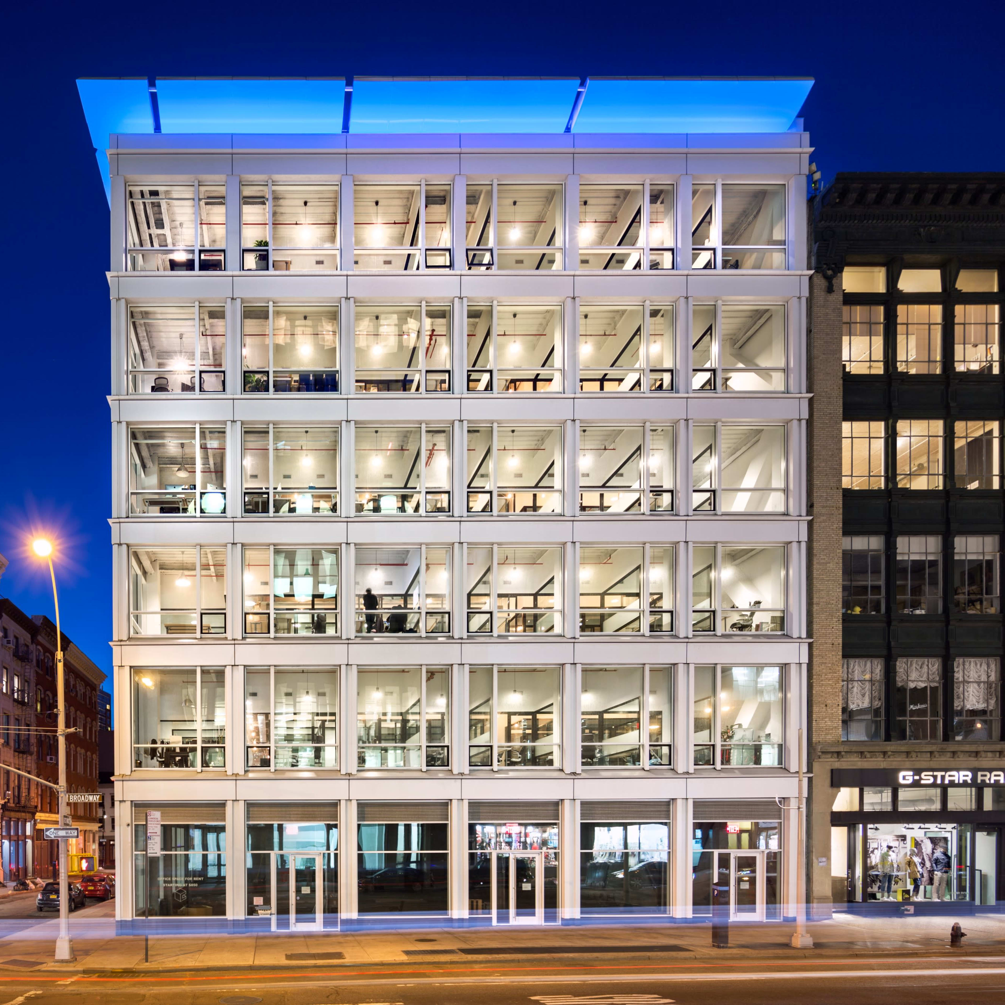 A modern glass-faced office building alongside historic brick buildings, illuminated at night with a blue-lit canopy.
