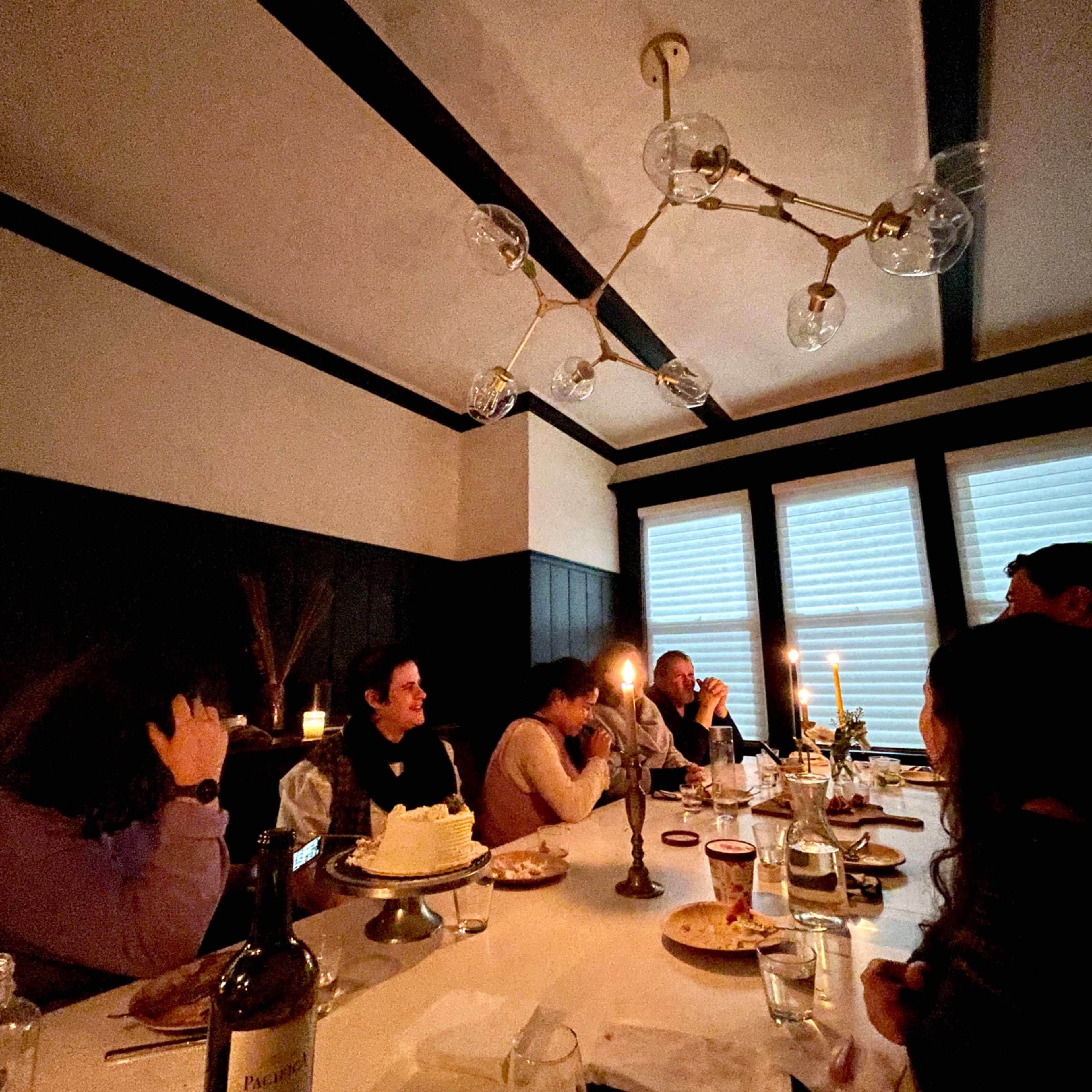A group of people sits around a long table adorned with food and candles, while dim lighting casts a warm glow on the scene.