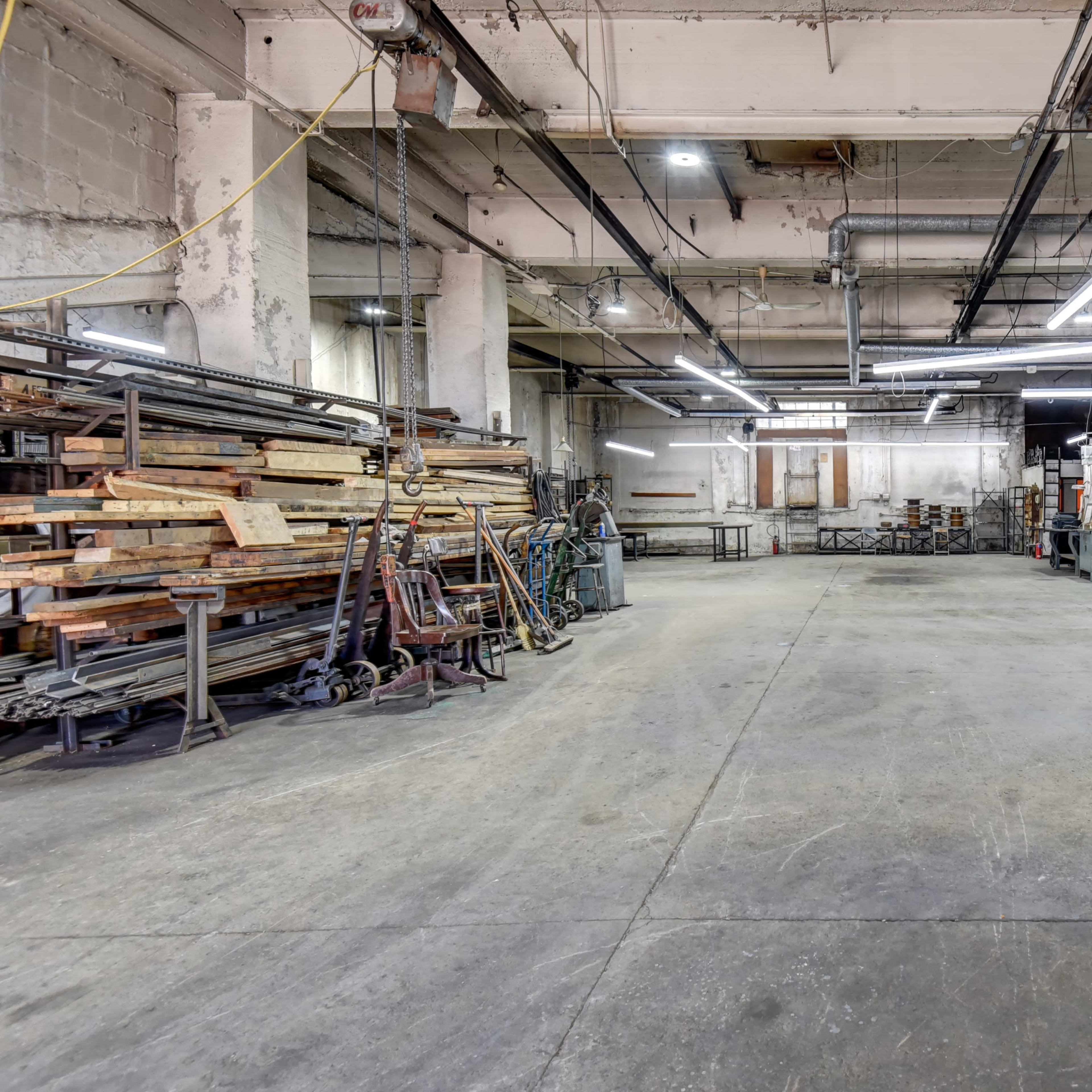 An empty industrial workshop features stacks of lumber, metalworking equipment, and overhead lighting.